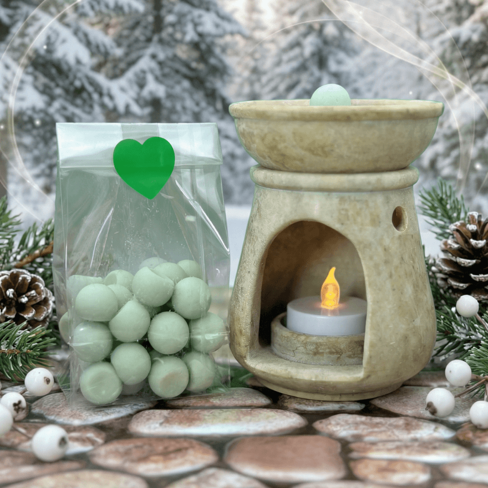 Pale green spherical wax melts in clear bag with heart sticker, displayed beside cream-colored ceramic wax warmer with lit tealight, arranged on wooden surface with snowy pine branches and pinecones.