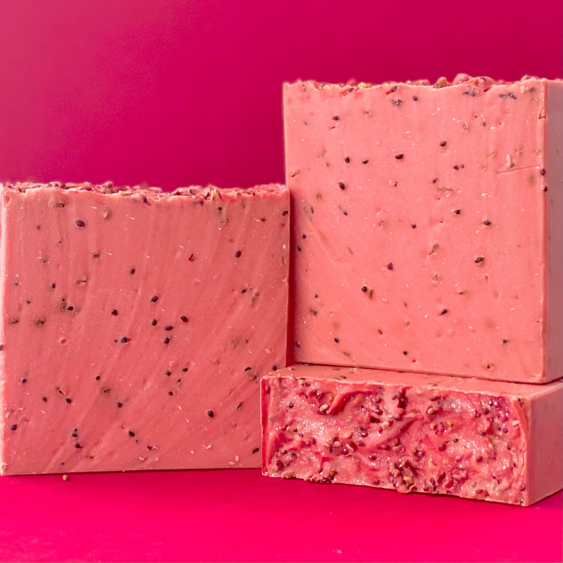 Three blocks of coral pink handmade soap bars with visible dark berry seeds throughout, displayed against a bright pink background. One bar is cut to show the speckled interior texture.