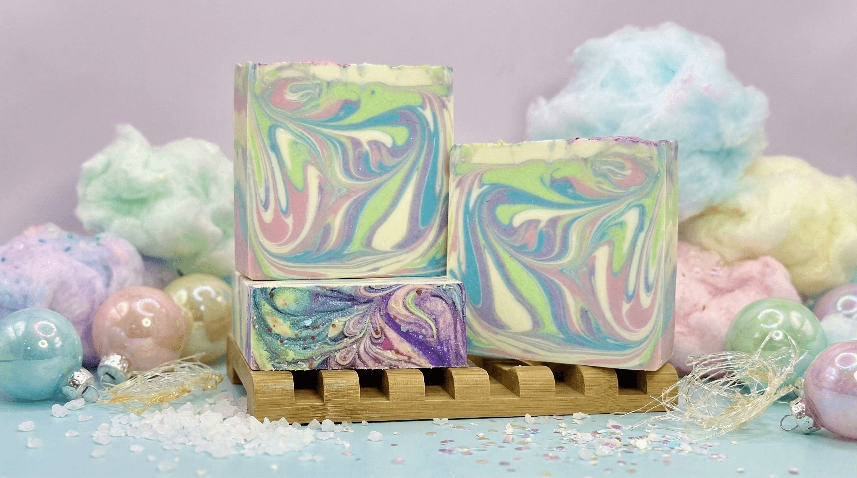 Three pastel swirled handmade soap bars in yellow, pink, green, and purple marble patterns displayed on a wooden soap dish with fluffy cotton candy props and iridescent ornaments.