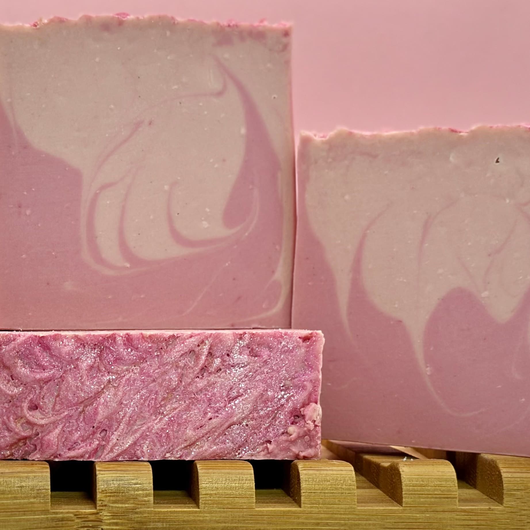 Three pink marbled soap bars on wooden slat rack against pink background. Bars show swirled pattern, with front bar displaying glittery pink surface texture and fine pumice exfoliation.