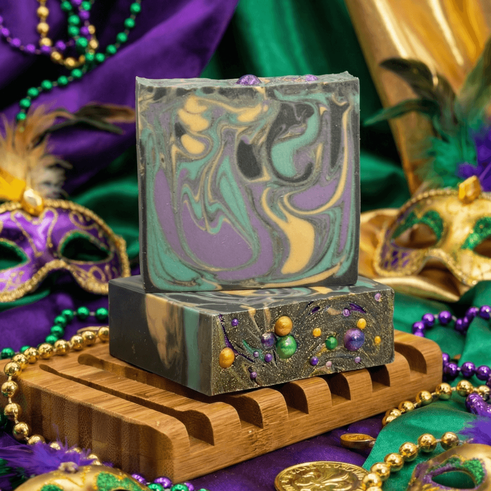 Two stacked handmade soap bars on a wooden soap dish with Mardi Gras decorations. Top bar features purple, green, and gold swirled pattern; bottom bar has glittery black base with embedded colored beads.