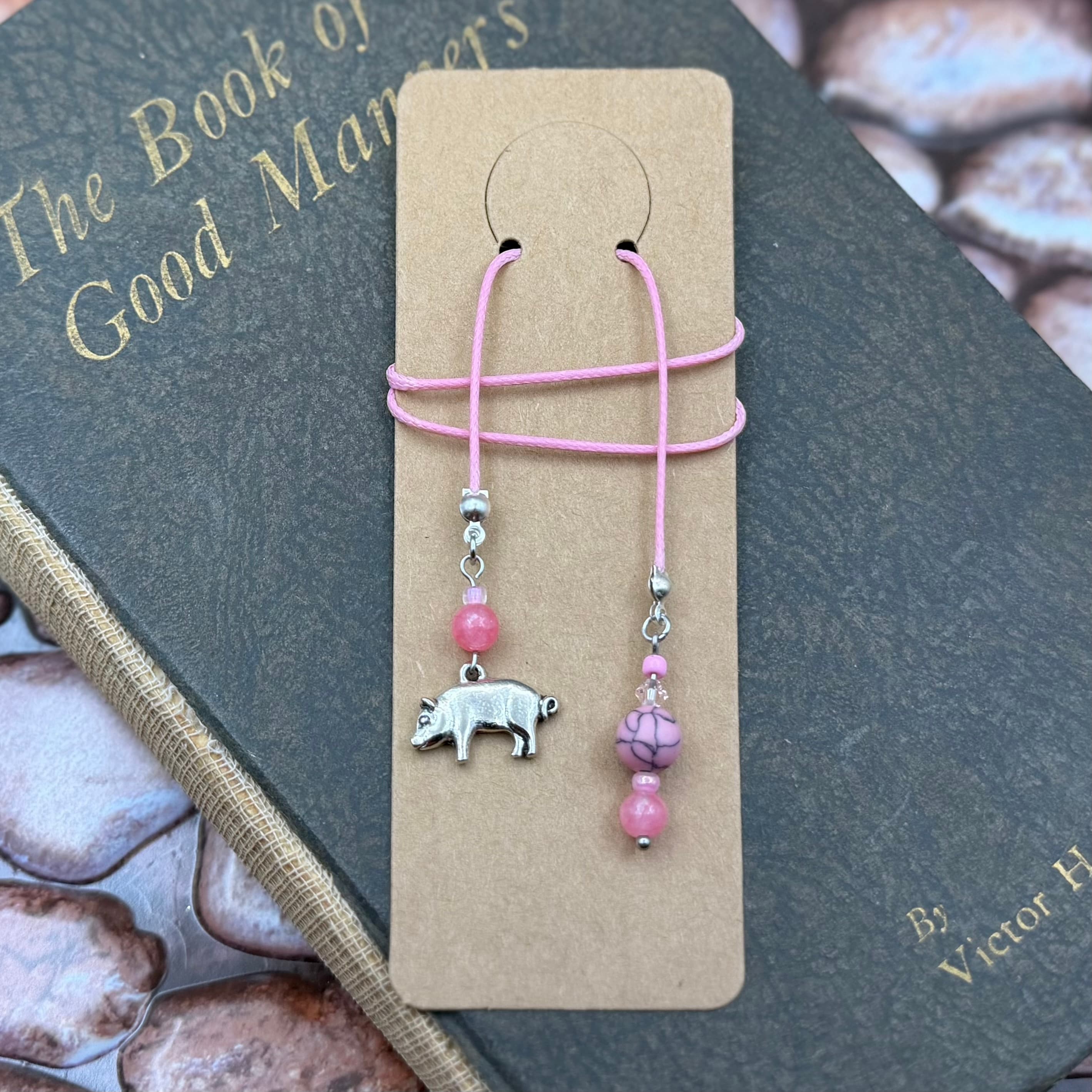 Handmade bookmark with pink cord and two beaded tassels displayed on kraft card over vintage book. One tassel features a silver pig charm, the other has pink and purple beads.