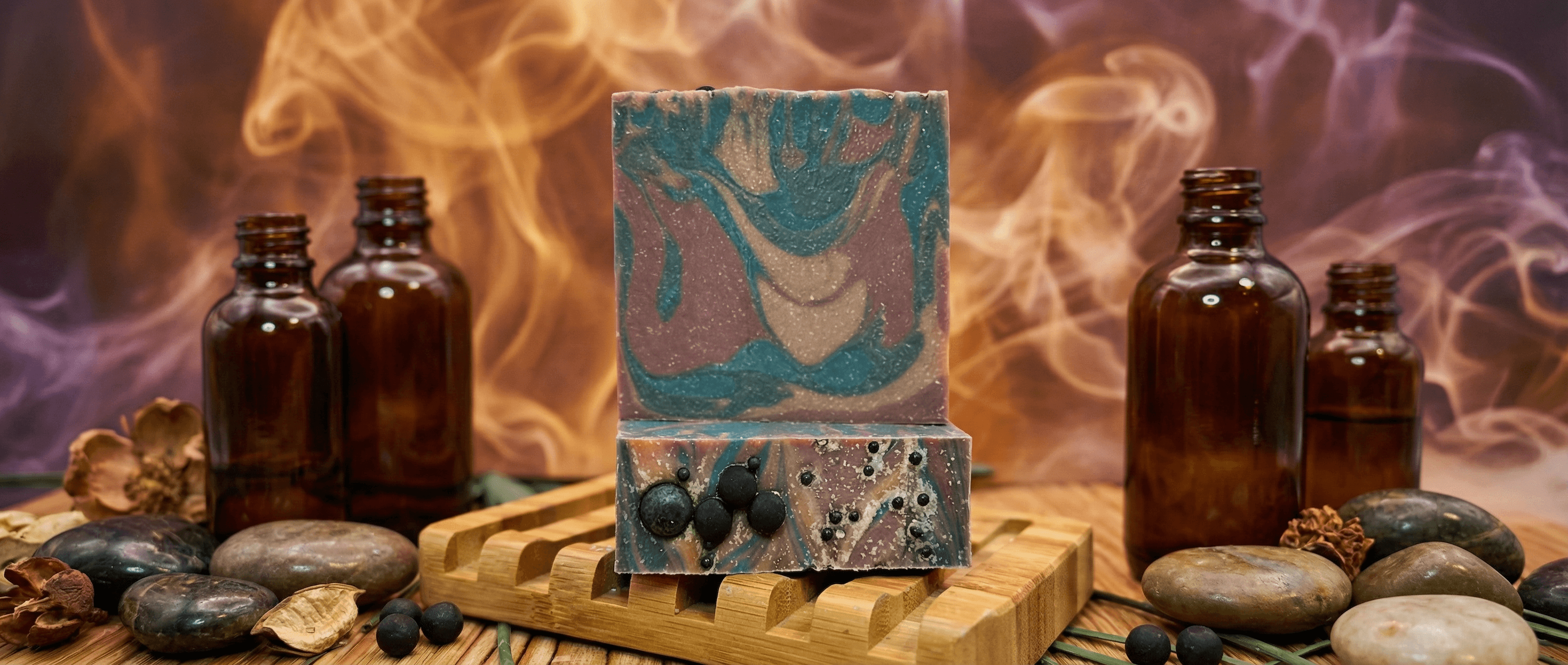 Two marbled soap bars stacked on a wooden dish, featuring swirled teal, mauve, and gold colors with visible black spherical botanicals embedded in the surface. Styled with smooth river stones and amber glass bottles.
