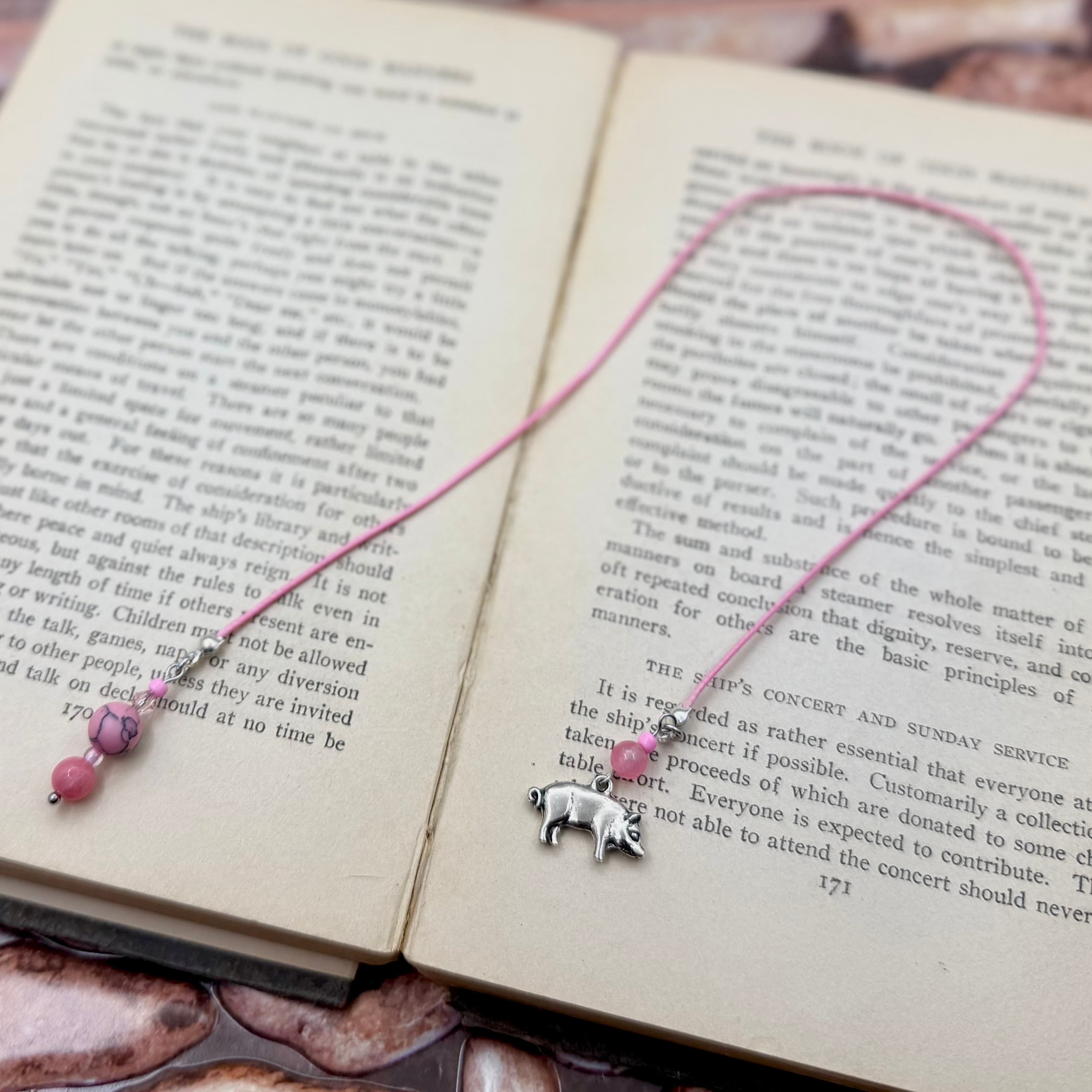 Pink cord bookmark with silver pig charm and pink beads laid across open book pages. The bookmark forms a heart shape displaying the full design and beaded details.