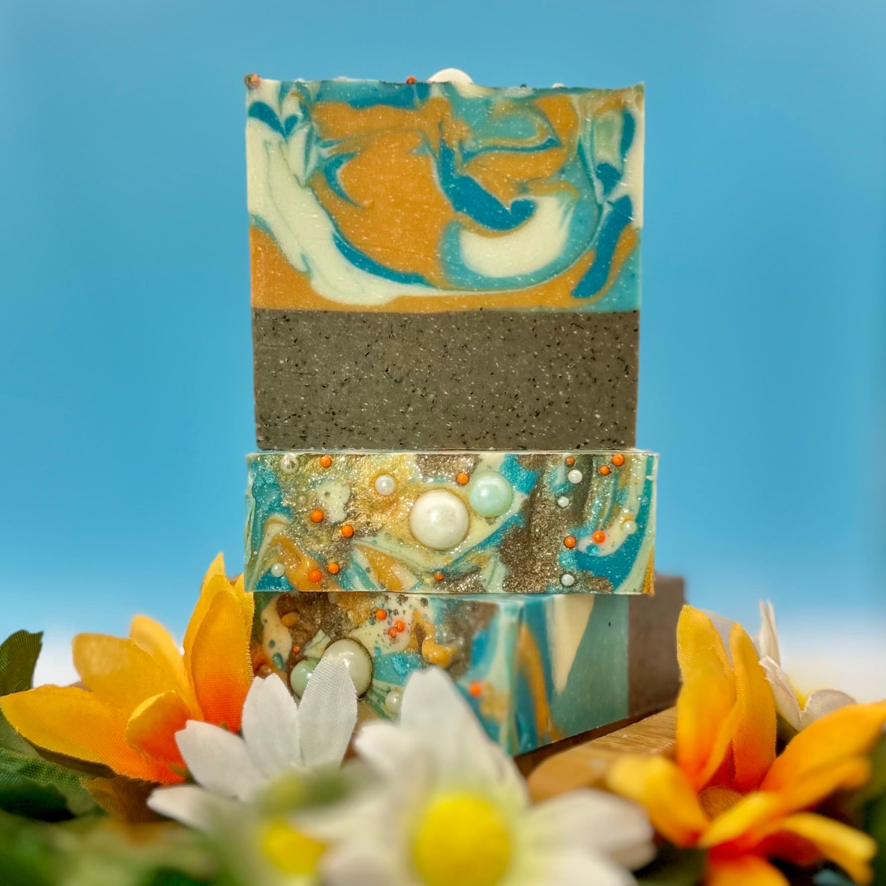 Three stacked bars of handmade soap with swirled turquoise, cream, and golden orange marbling on top, dark gray exfoliating layer below, decorated with white and orange pearl embeds, surrounded by yellow and white flowers.