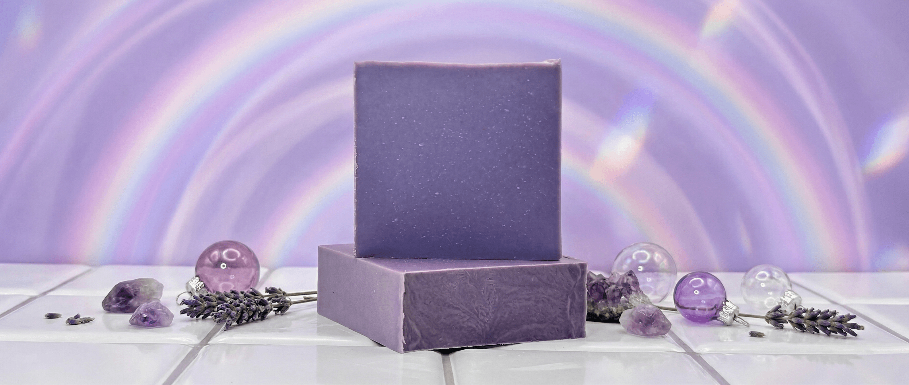 Two purple lavender soap bars with textured surface displayed with amethyst crystals, glass orbs, and dried lavender sprigs on white tile against swirling purple background.