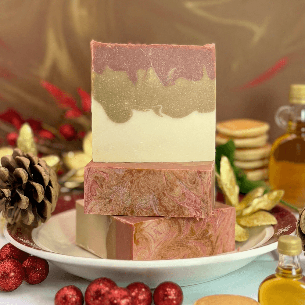 Three bars of handcrafted soap stacked on a white pedestal plate with holiday decor. Top bar shows layered design in pink, tan, and cream. Bottom bars feature pink and tan swirls.