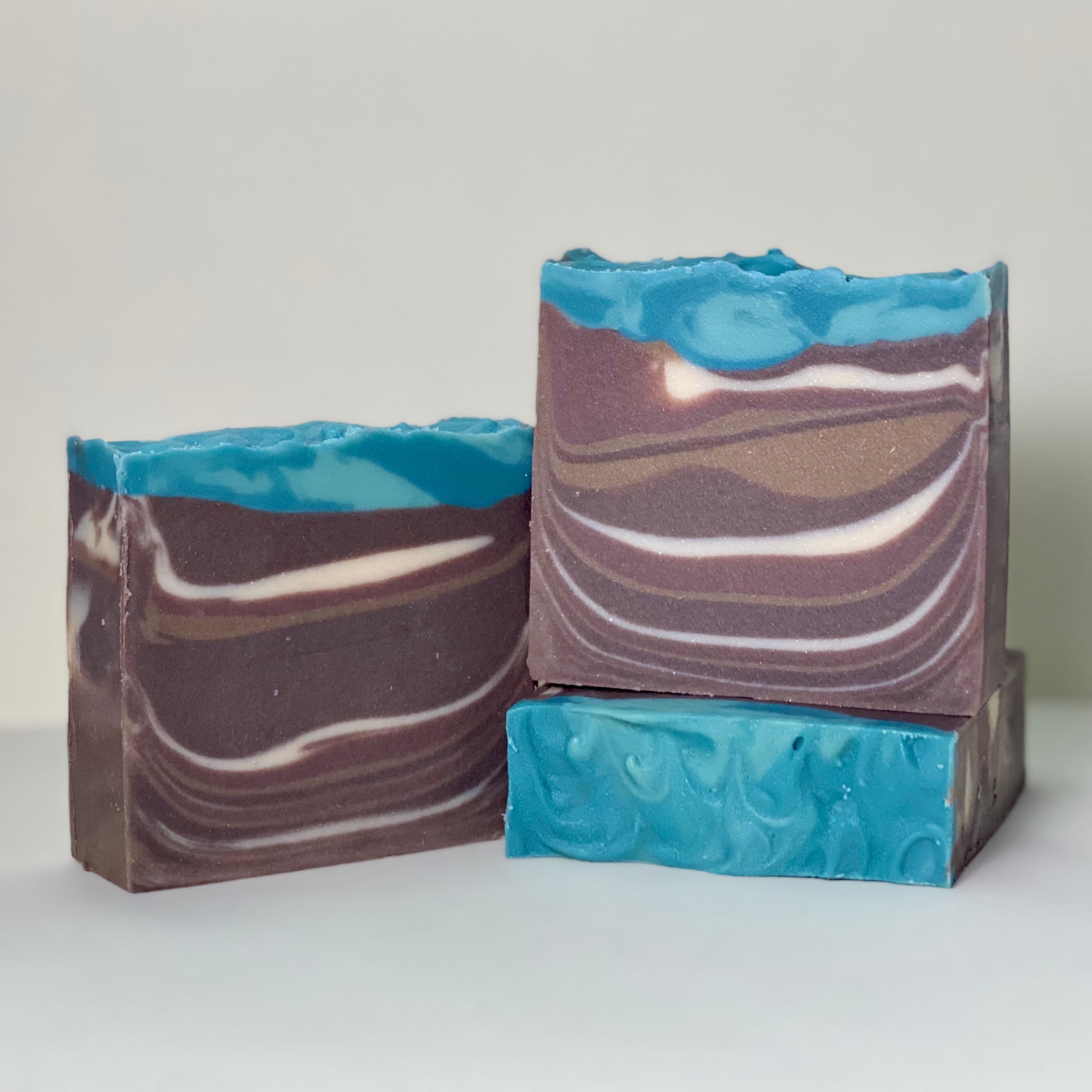 Three rectangular bars with swirled layers of deep purple-brown and white, topped with bright turquoise. The soap has flowing, wavy patterns throughout creating an artistic marbled effect.