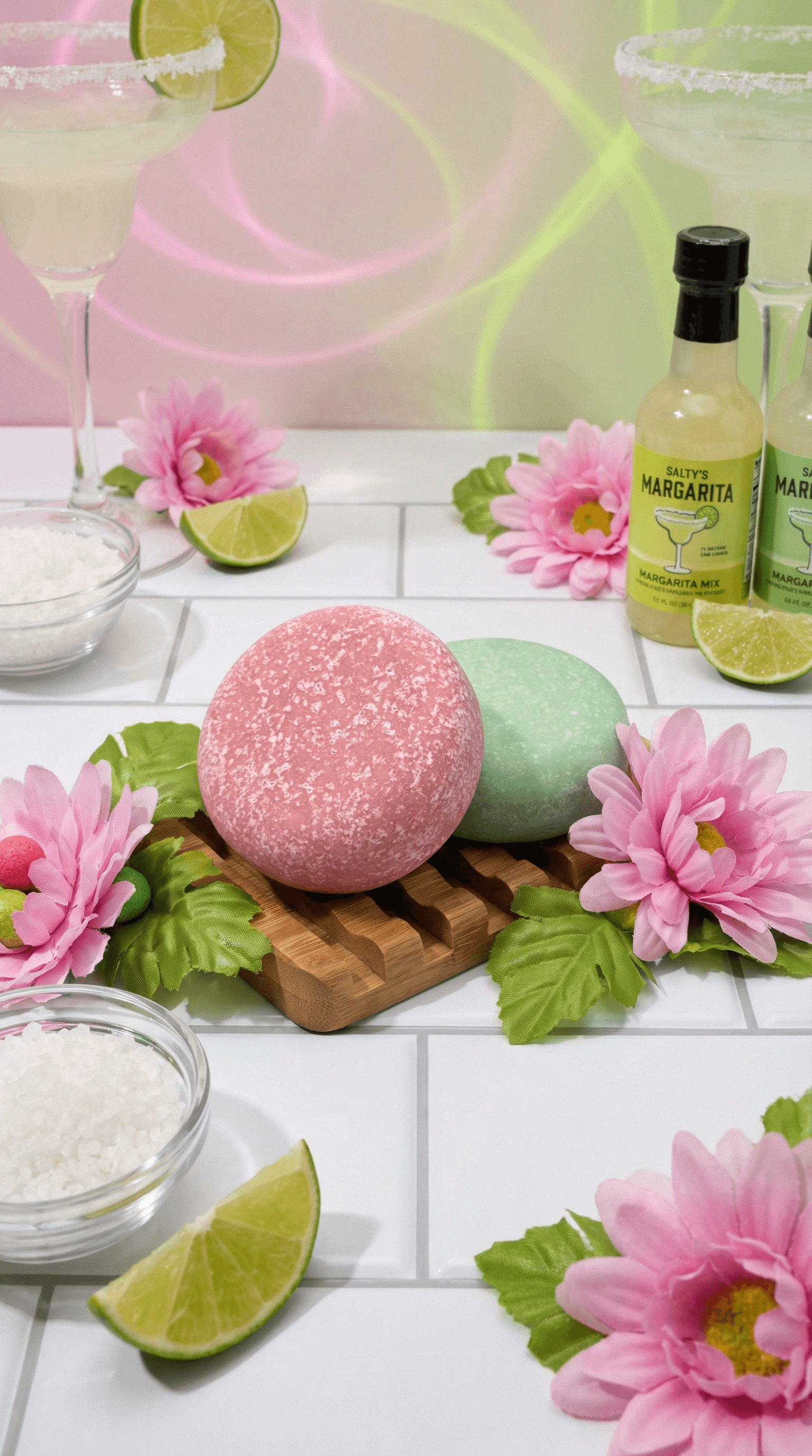 Two round textured sea salt scrub bars on a wooden tray, one coral pink and one mint green, surrounded by pink flowers, lime wedges, salt, and margarita glasses with yellow-green bottle in background.