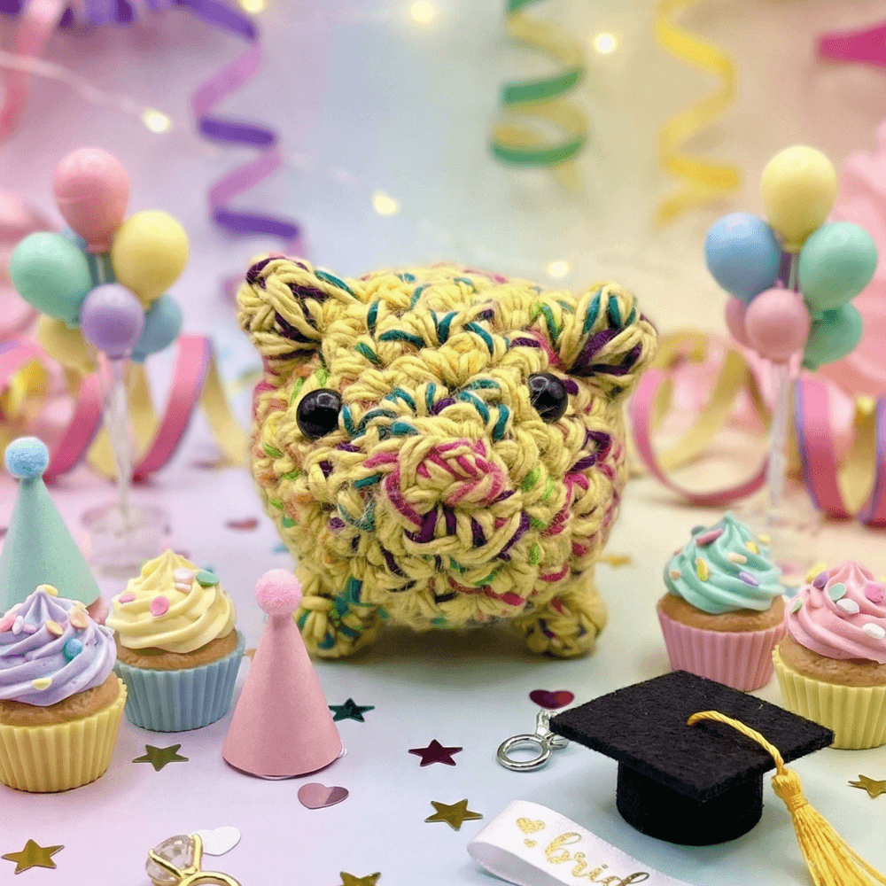 Yellow crocheted pig-shaped sachet with colorful rainbow flecks, black bead eyes, and snout detail, displayed among pastel cupcakes, balloons, and party decorations.