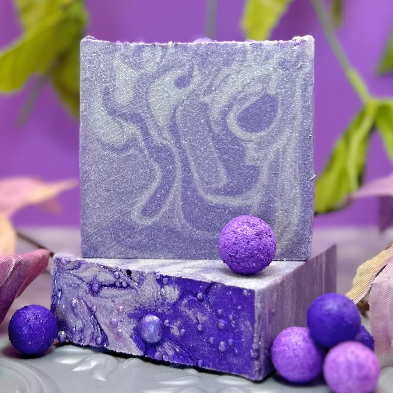 Two purple handmade soap bars stacked with swirled patterns and decorative purple spheres scattered around. The top bar has a smooth, frosted appearance while the bottom shows marbled bubbles.
