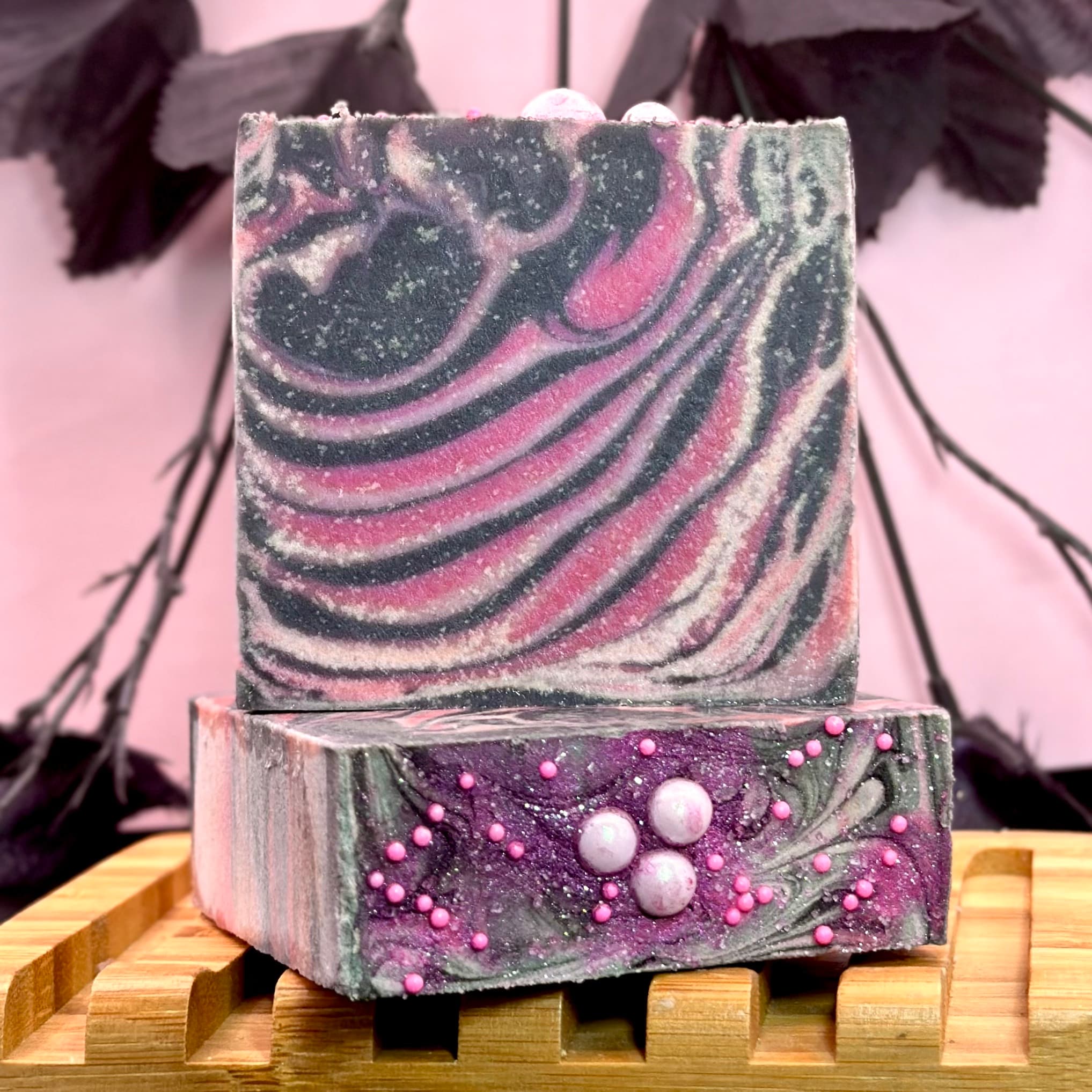 Two stacked handcrafted soap bars on a wooden soap dish. Top bar has swirled charcoal and pink marbling. Bottom bar features purple glitter with pink and white decorative pearls.