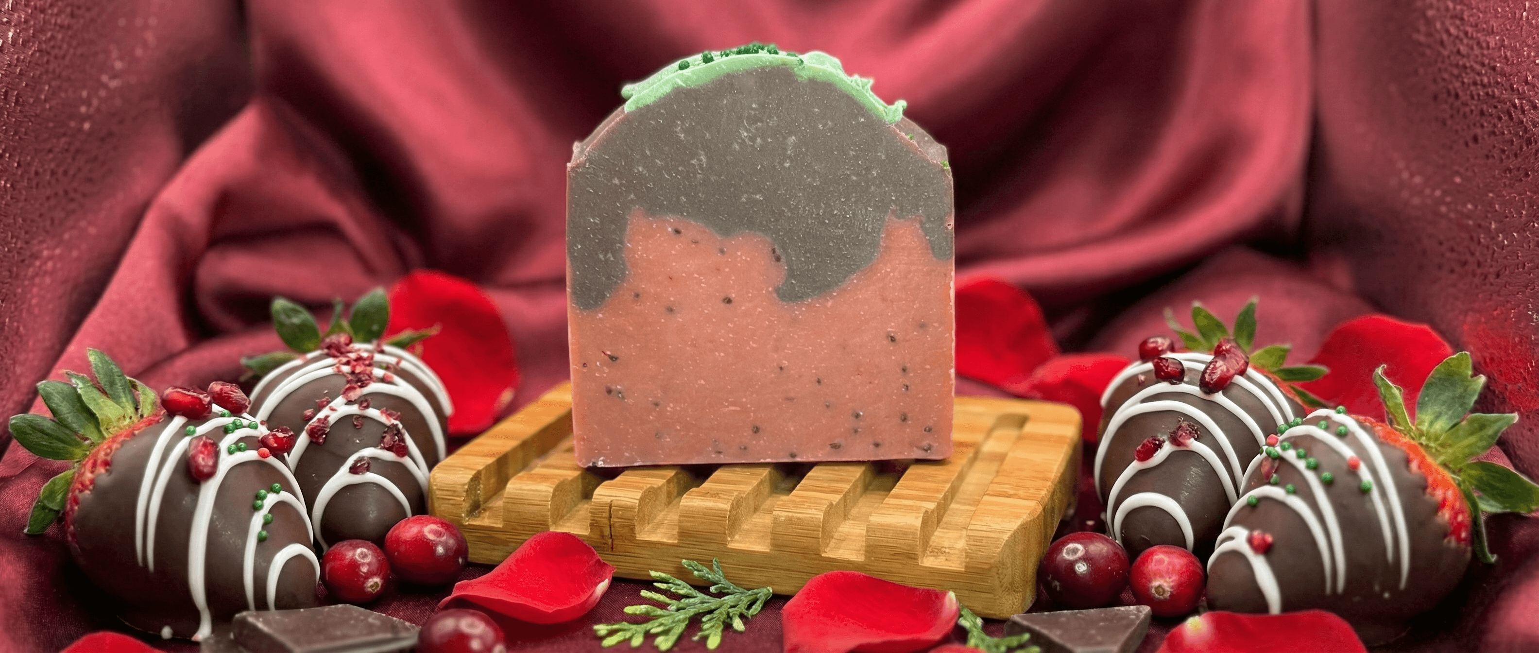 Handmade soap bar with pink bottom layer speckled with dark seeds, gray top layer, and green frosting-style piped edge, displayed on wooden soap dish with chocolate-covered strawberries and festive botanicals.