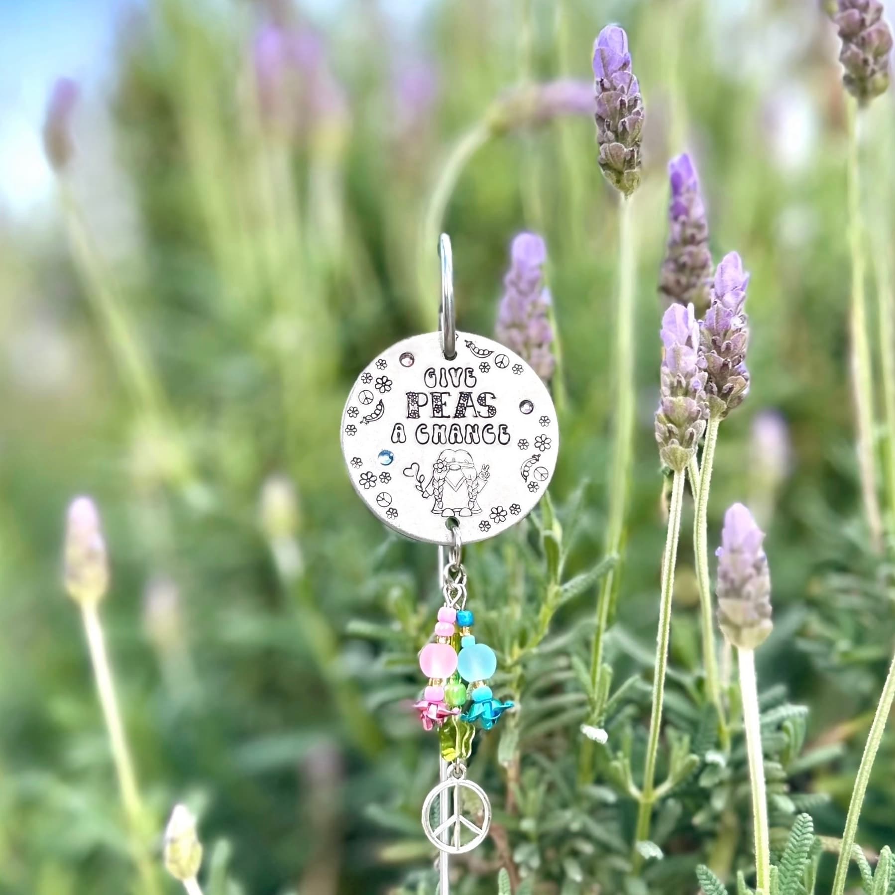 Round silver metal disc stamped with peace symbol message and gnome illustration, adorned with colorful beaded charm featuring pink, blue and green glass beads with peace sign charm, displayed among purple lavender flowers in garden setting.