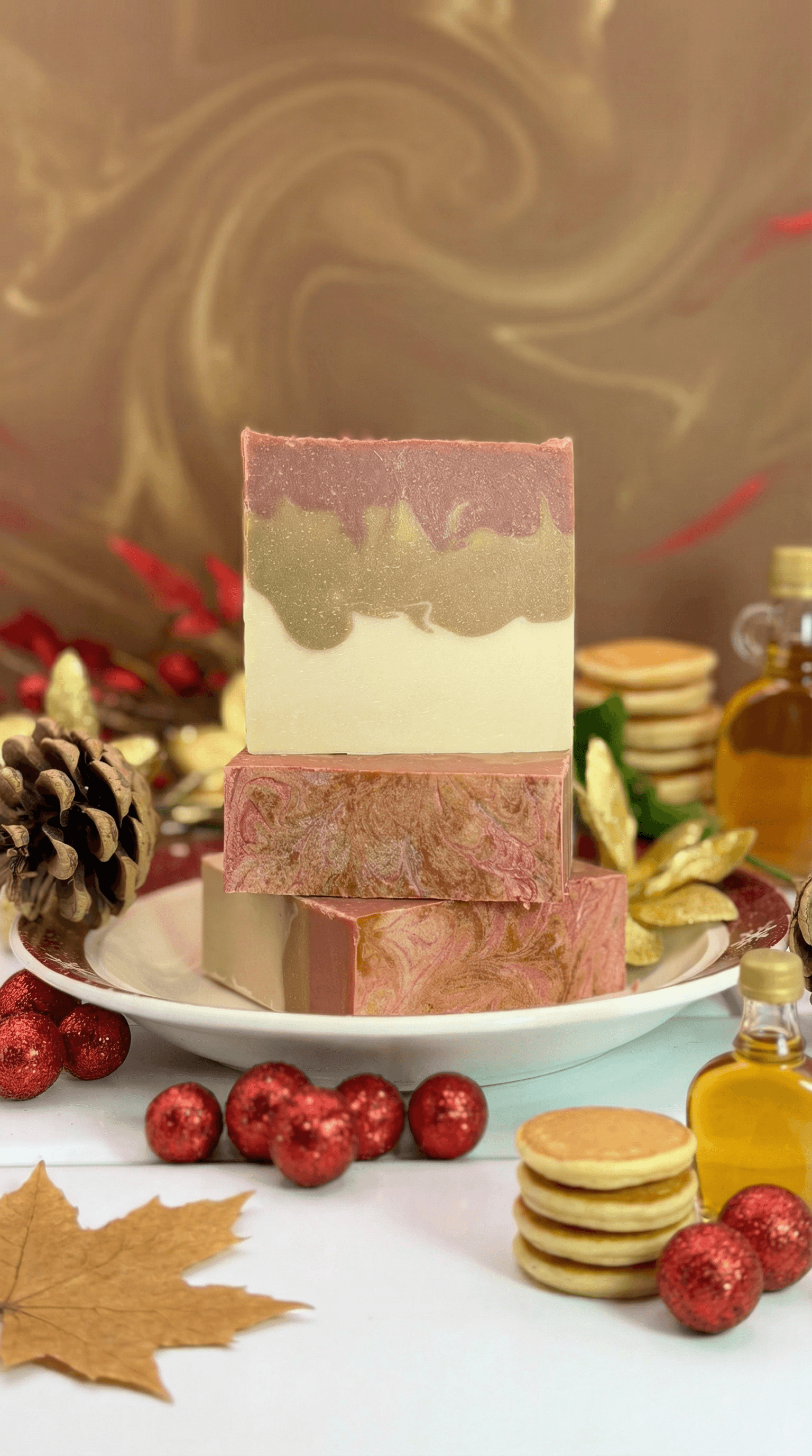 Stack of handcrafted soap bars on white pedestal plate with autumn leaves, pinecones, red ornaments, and small bottles of golden syrup. Top bar features layered design in pink, olive green, and cream with wavy edges.