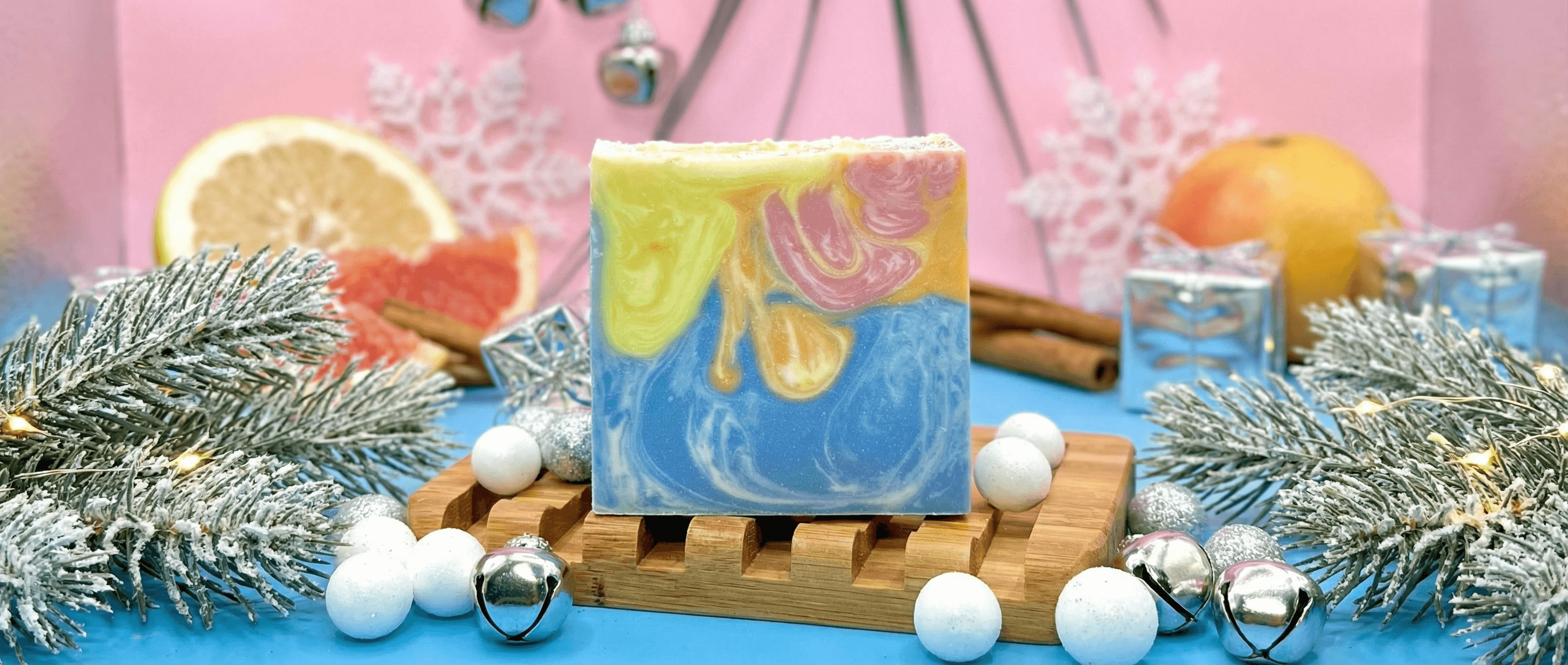 Handcrafted soap bar with swirled blue base and vibrant yellow, pink, and orange marbled top, displayed on wooden soap dish surrounded by frosted evergreen branches, citrus slices, cinnamon sticks, and silver ornaments.