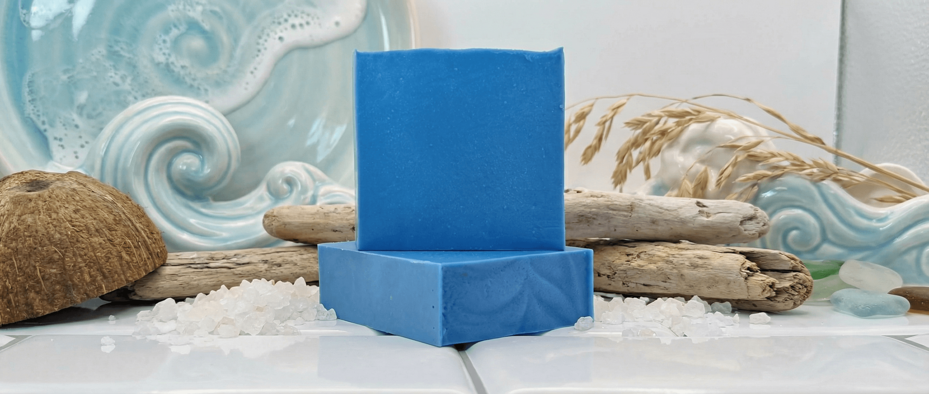 Two stacked blue handmade soap bars with smooth matte finish, displayed on white salt crystals with driftwood, coconut shell, dried wheat, and ocean wave artwork backdrop.