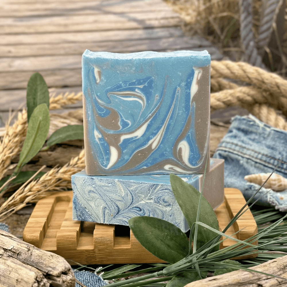 Two blue and cream swirled handmade soap bars stacked on a wooden soap dish, surrounded by wheat stalks, green leaves, rope, driftwood, and denim fabric in a coastal-themed styling.