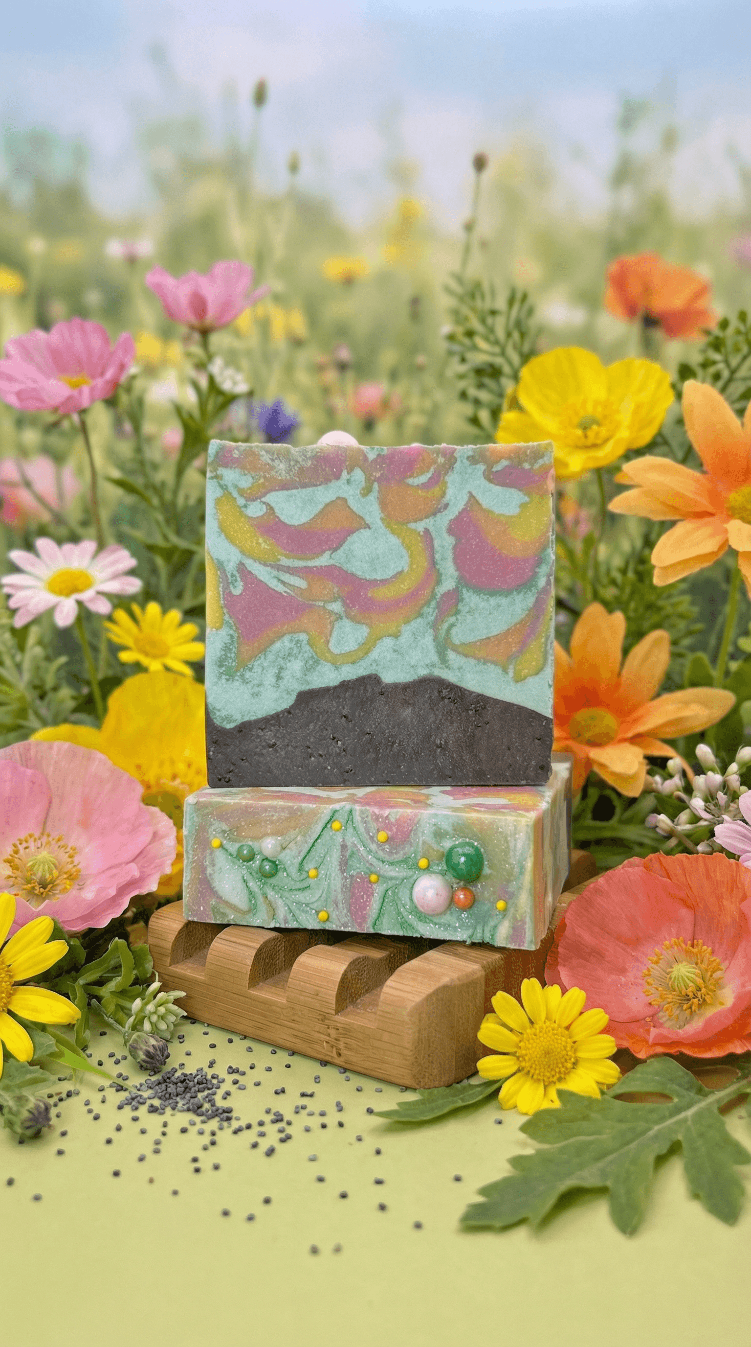 Two stacked handcrafted soap bars on a bamboo dish surrounded by wildflowers. Top bar features swirled teal, pink, and gold with dark brown base. Bottom bar has green swirls with embedded colorful sphere embeds.
