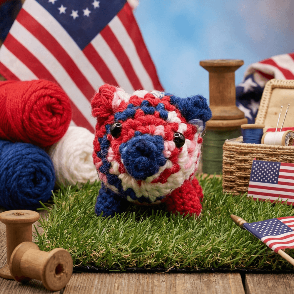 Hand-crocheted patriotic piggy sachet in red, white, and blue yarn with button eyes, displayed on artificial grass with wooden spools, yarn balls, and American flags in background.