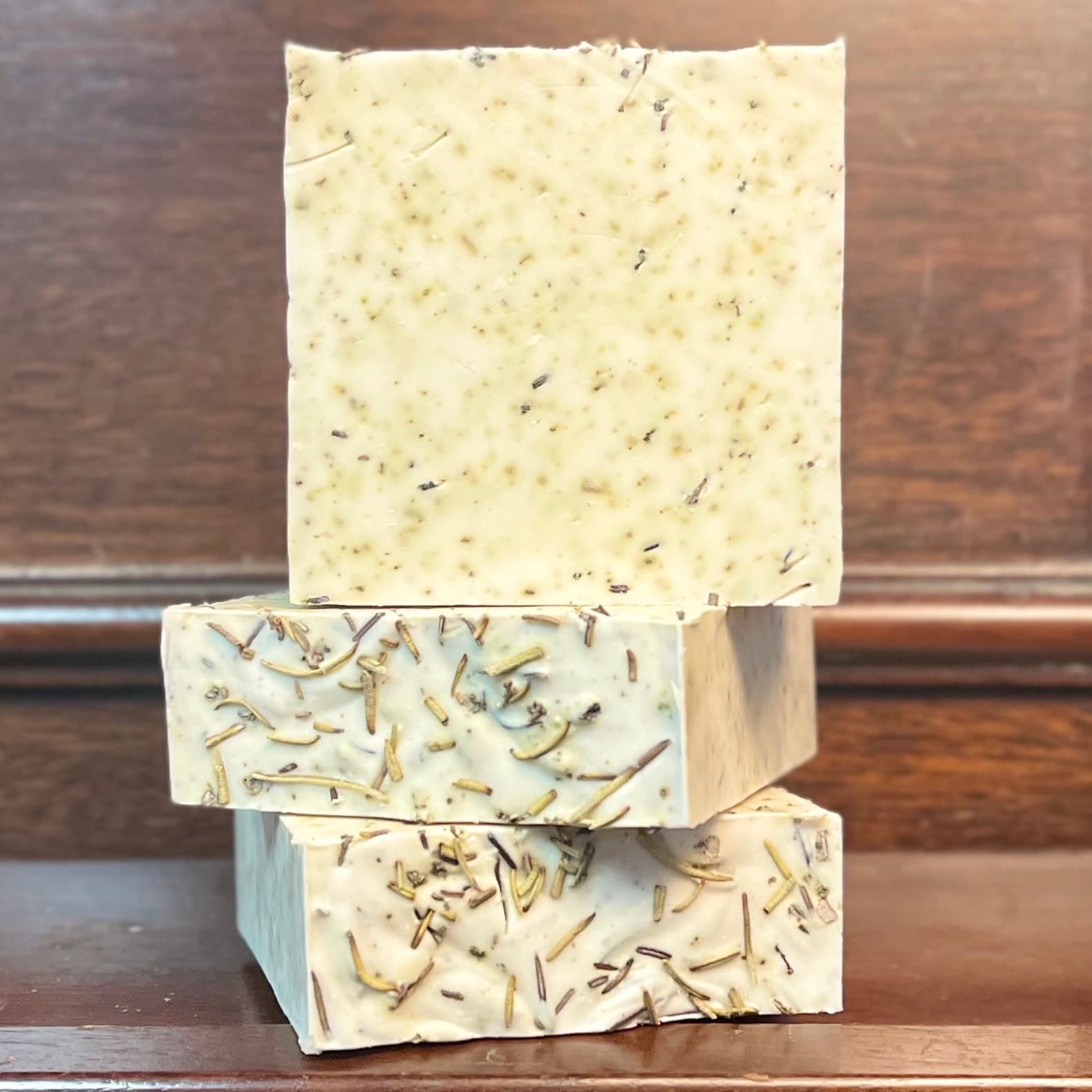 Three stacked bars of cream-colored handmade soap with visible sage and rosemary herbs embedded throughout, displayed on a wooden surface against a rustic background.