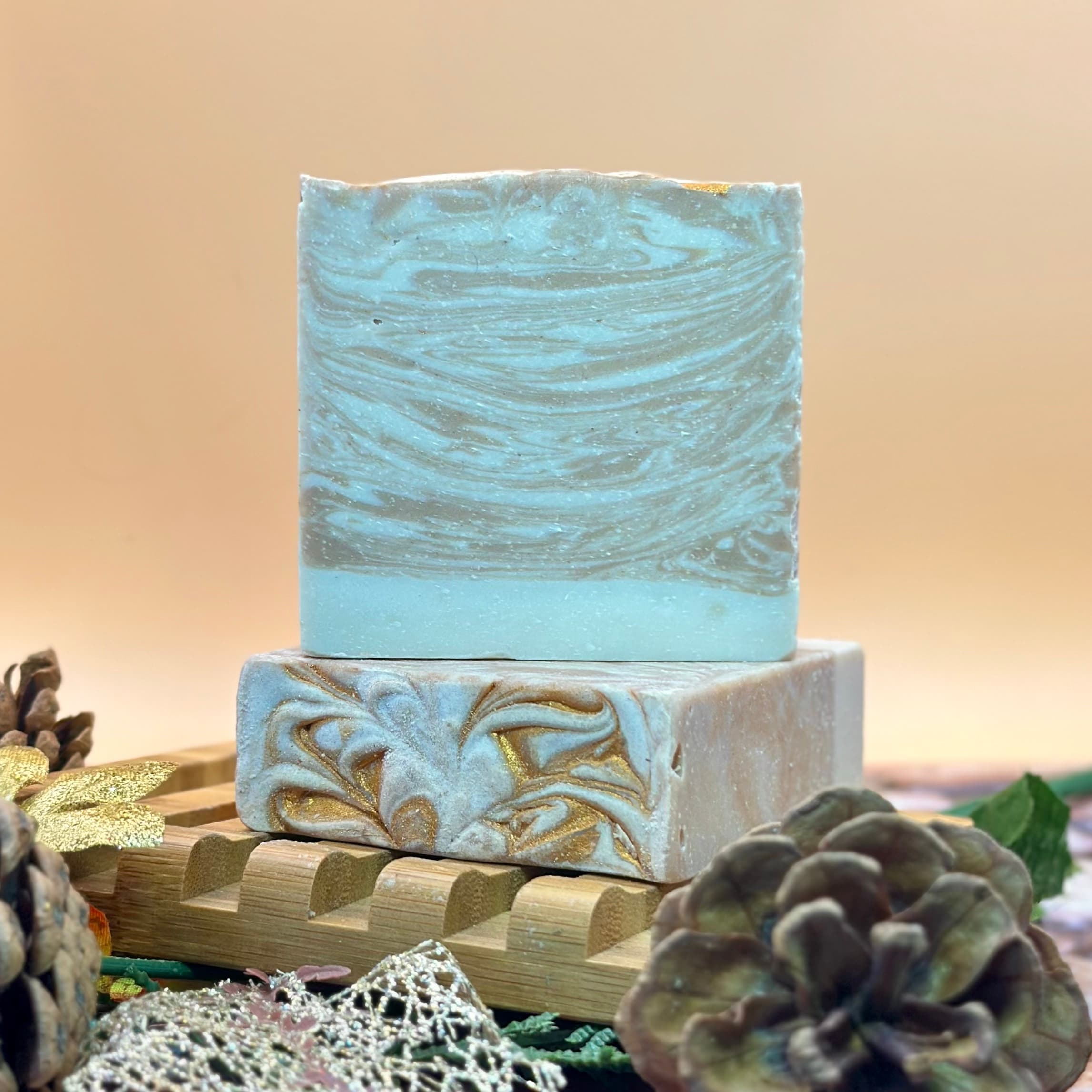 Two stacked artisan soap bars in pale blue with gold swirled designs, displayed on a bamboo tray with pine cones and succulent plants against a neutral background.