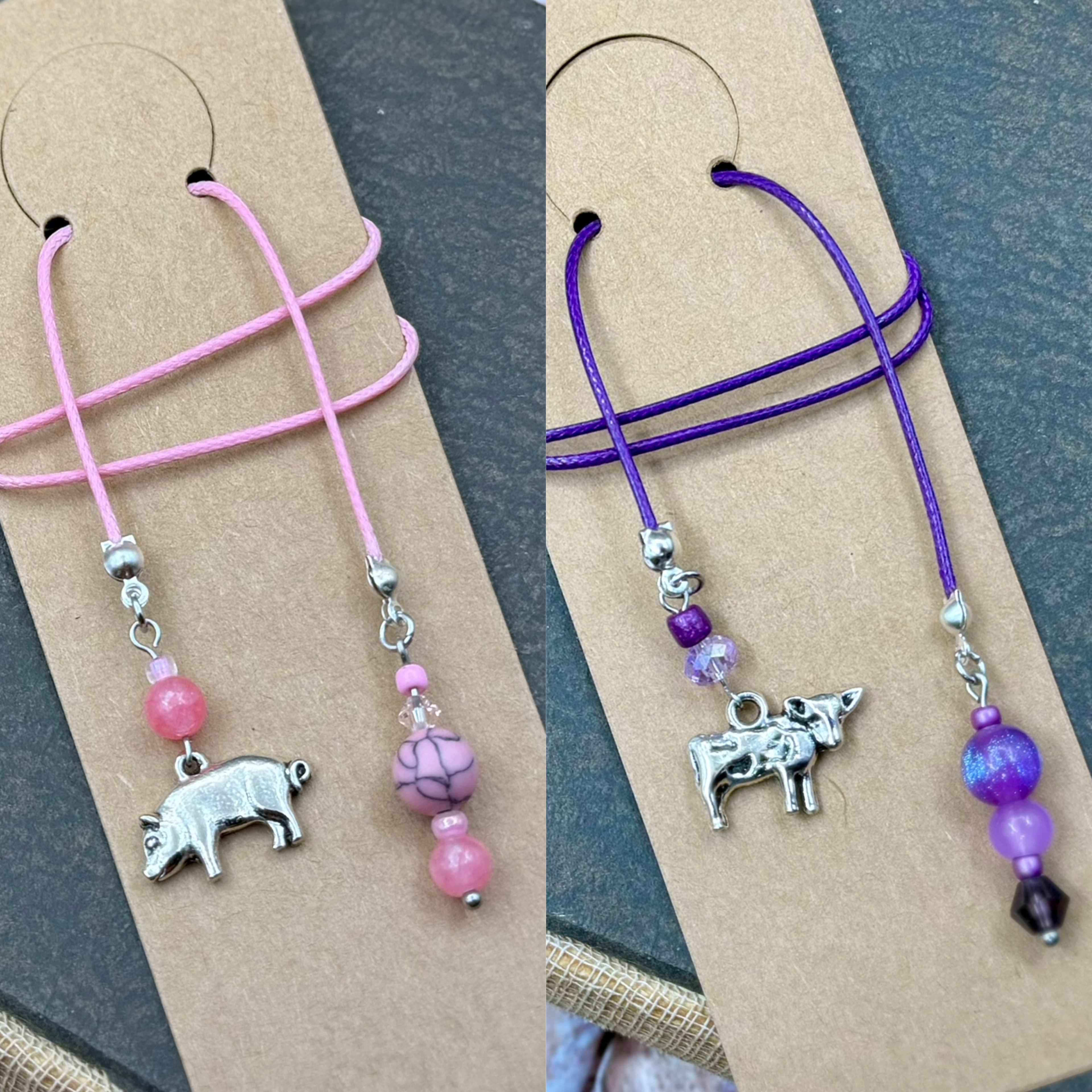 Two handmade bookmarks with pig and cow silver charms. One features pink cord and beads, the other purple cord and beads. Both displayed on tan cardstock tags.
