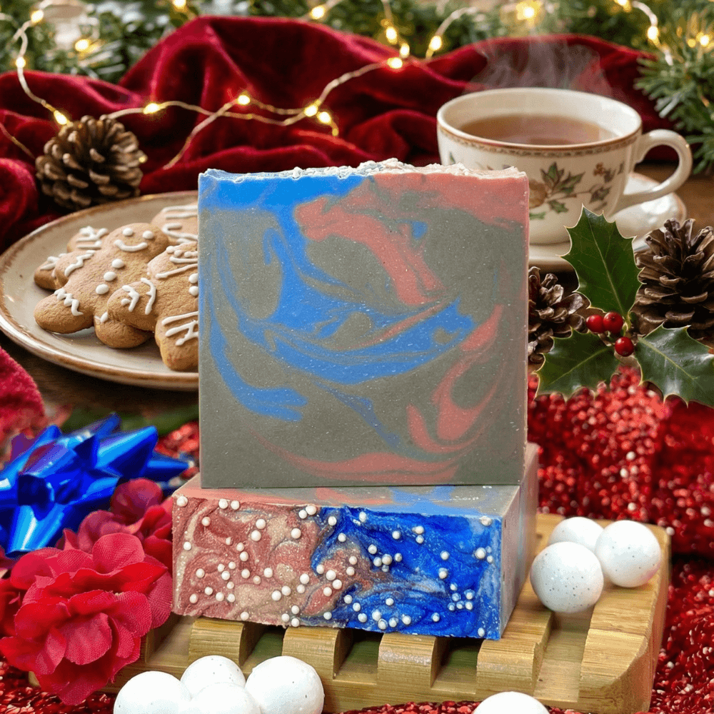 Two stacked handmade soap bars with swirled blue, coral pink, and sage green marbling, displayed on a wooden soap dish. Surrounded by gingerbread cookies, tea, pine cones, and holiday decorations.