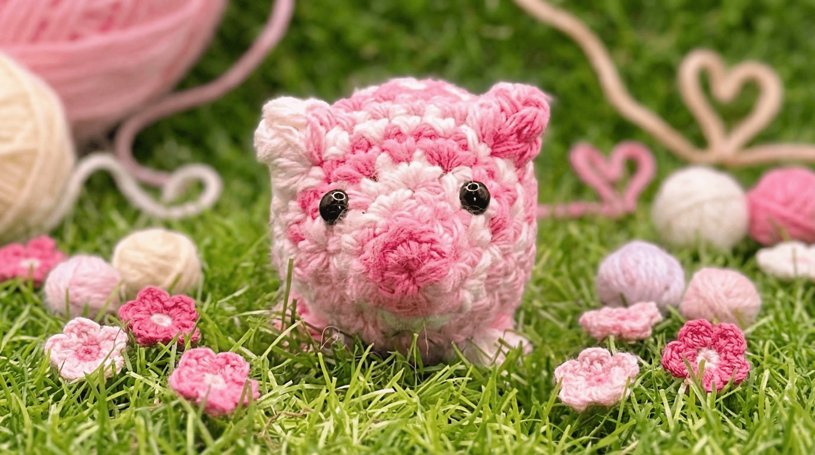 Hand-crocheted pink and white pig-shaped sachet on artificial grass, surrounded by small crocheted flowers and yarn balls. The pig has black bead eyes and variegated pink stitching.
