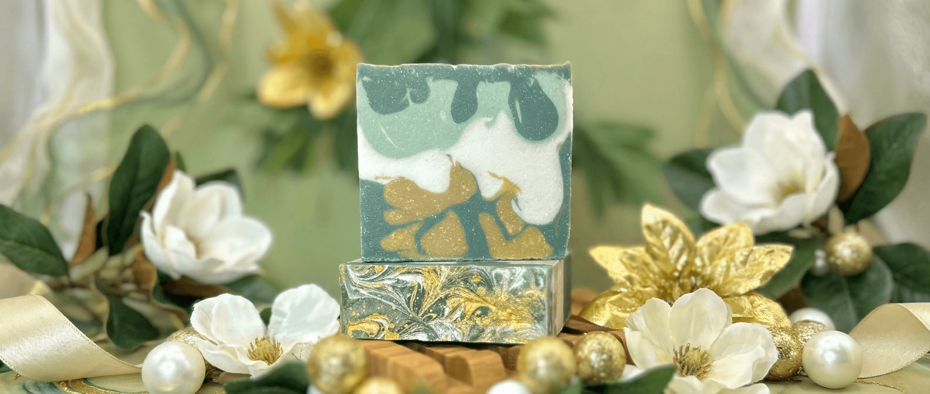 Two stacked handcrafted soap bars with swirled patterns in sage green, white, and metallic gold, displayed with white flowers, golden leaves, and pearl ornaments.