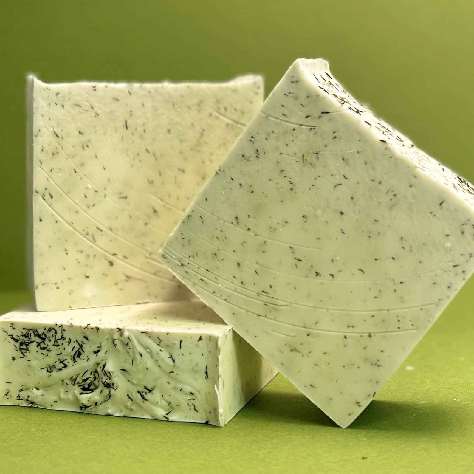 Three bars of pale green handmade soap with dark herb flecks throughout, displayed stacked against a matching green background. The bars show swirled texture on their surface.