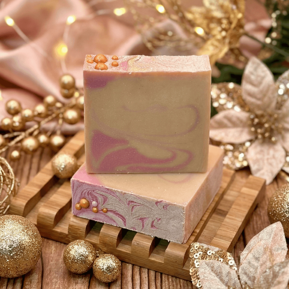 Two stacked bars of handmade soap in cream and dusty rose swirls, topped with gold biodegradable glitter and pearl accents, displayed on a wooden soap dish with festive gold holiday decorations.
