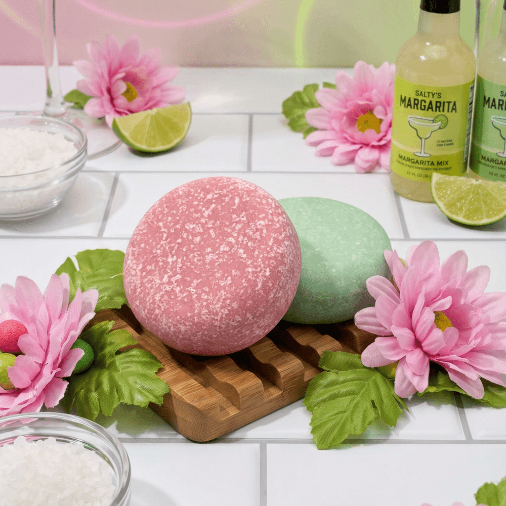 Two round sea salt scrub bars on a wooden tray, one coral pink with sparkly texture and one soft mint green, surrounded by pink flowers, lime slices, and margarita mix bottles.