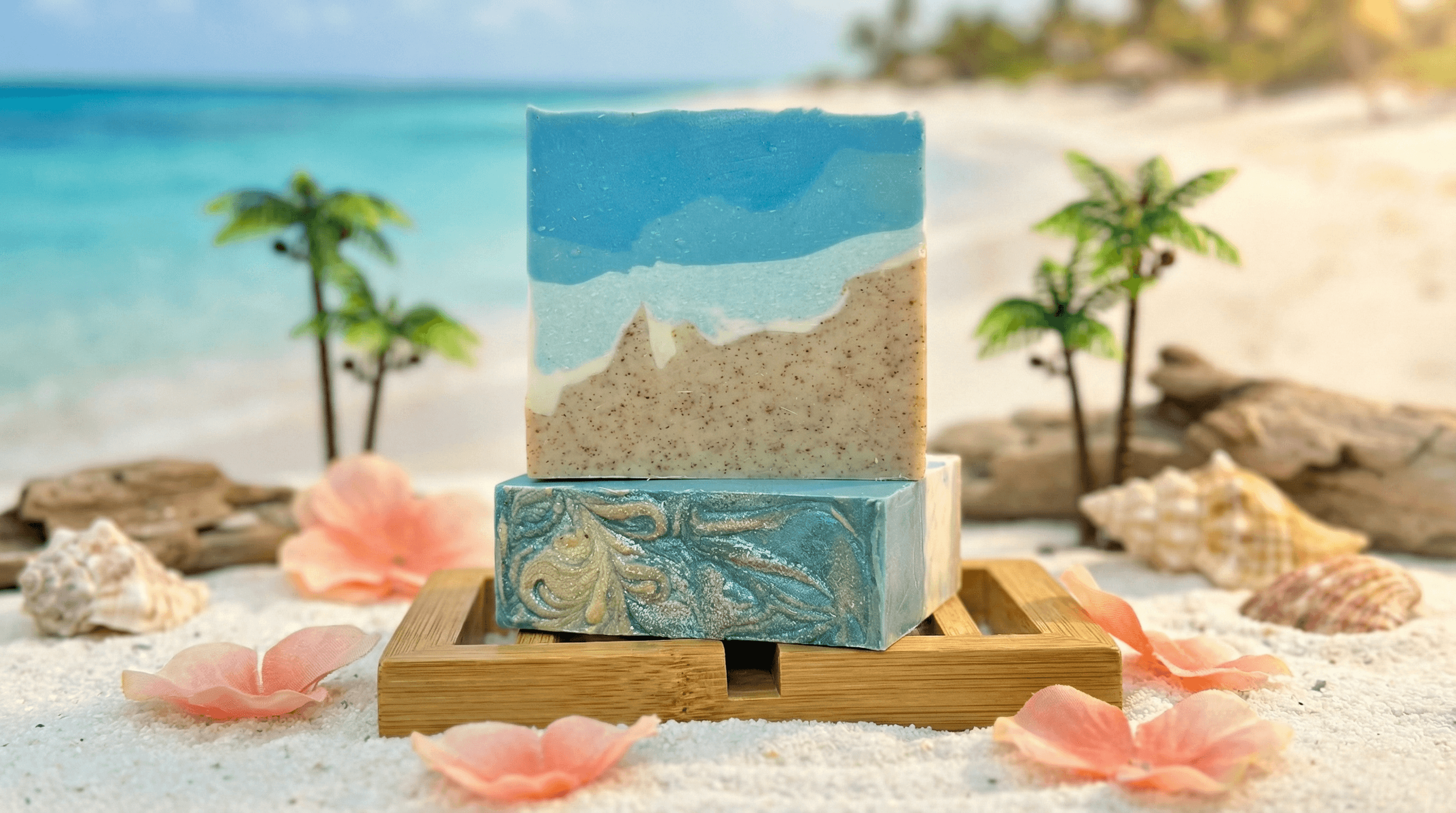 Two stacked handcrafted soap bars on bamboo dish with beach background. Top bar shows layered ocean waves in blue and tan sand with speckles. Bottom bar features swirled blue and gold marble pattern.