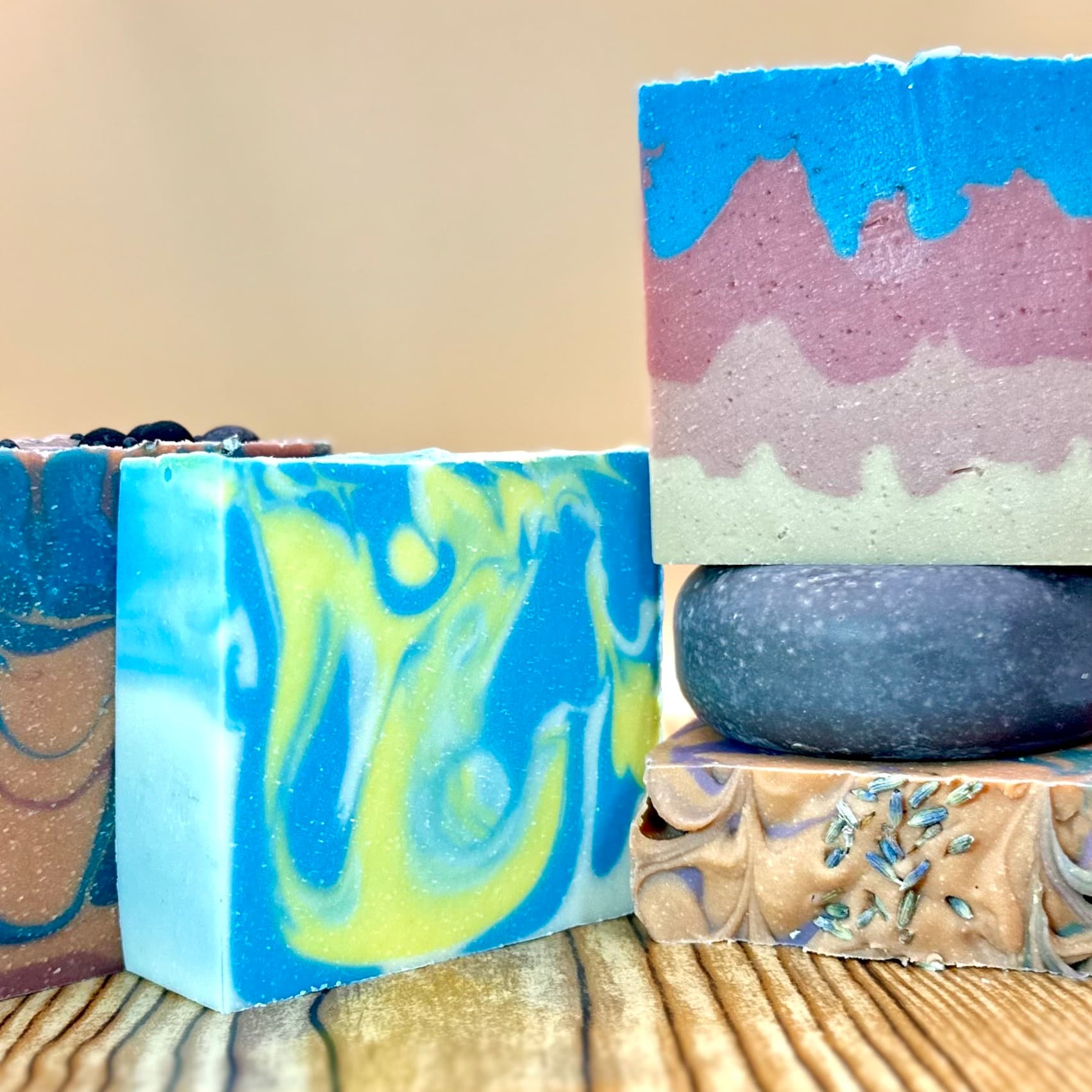 Five handcrafted soap bars arranged on wood: swirled blue-yellow marbled bar, layered purple-pink-mint bar, dark charcoal round bar, and tan bar with dried lavender buds on top.