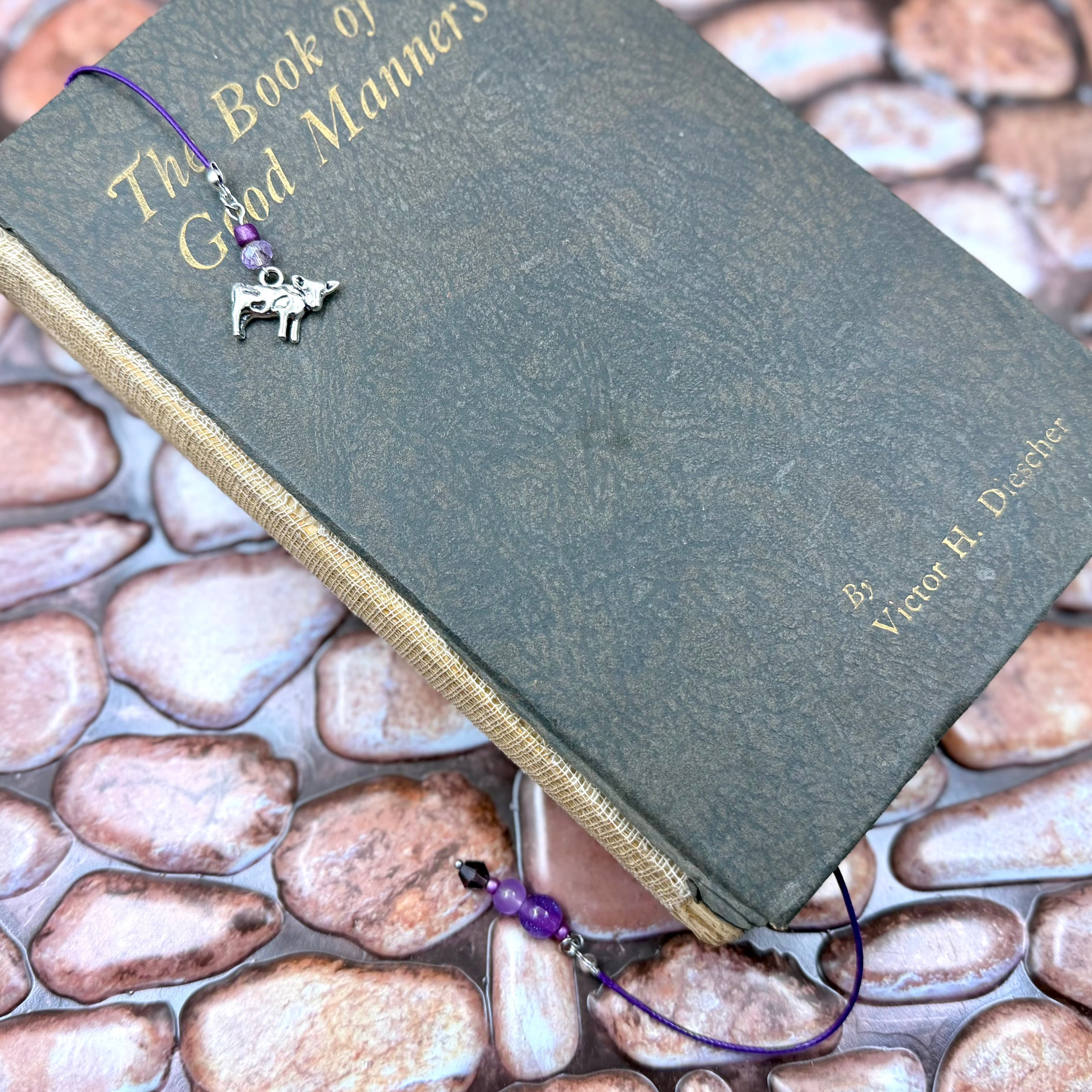 Dark gray book titled 'The Book of Good Manners' by Victor H. Diecher with purple cord bookmark featuring silver cow charm and purple beads, resting on decorative stones.