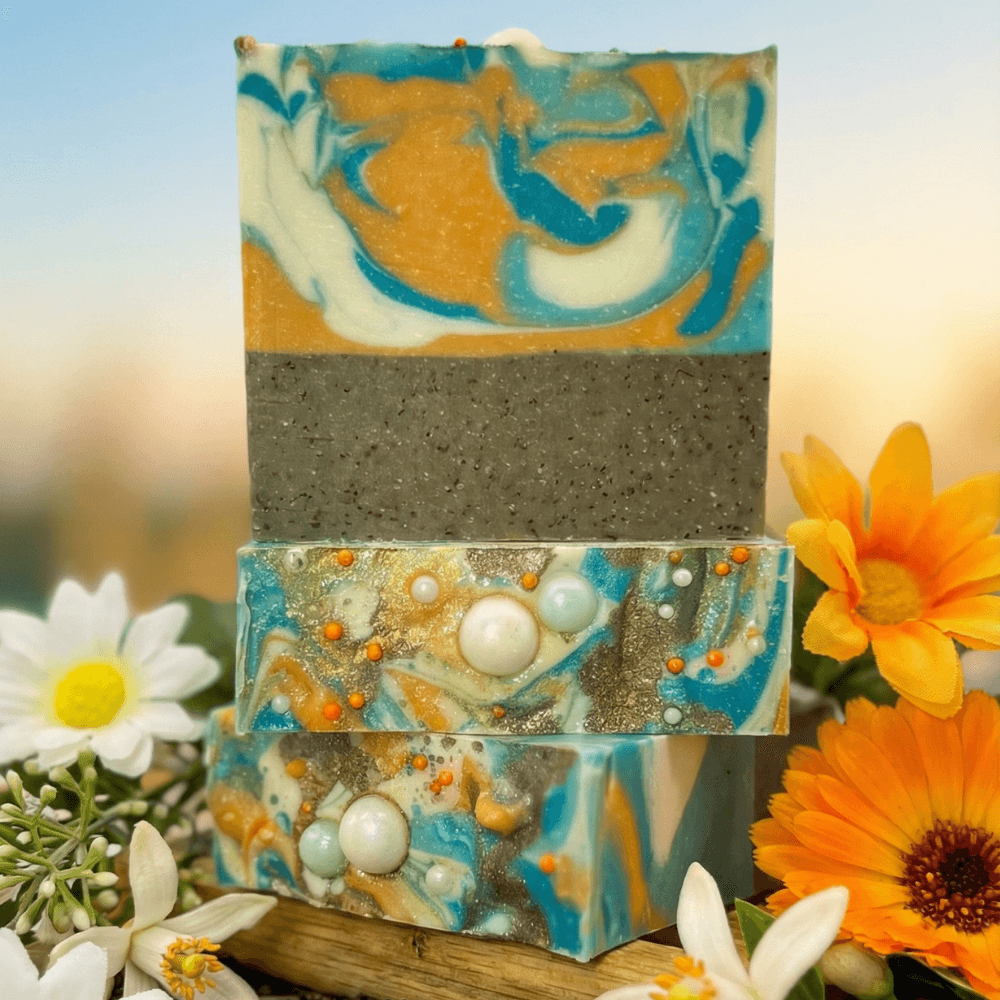 Three stacked handmade soap bars with swirled orange, turquoise, and cream top layer. Gray speckled bottom layer. White and orange decorative beads embedded. Displayed with daisies and marigolds.