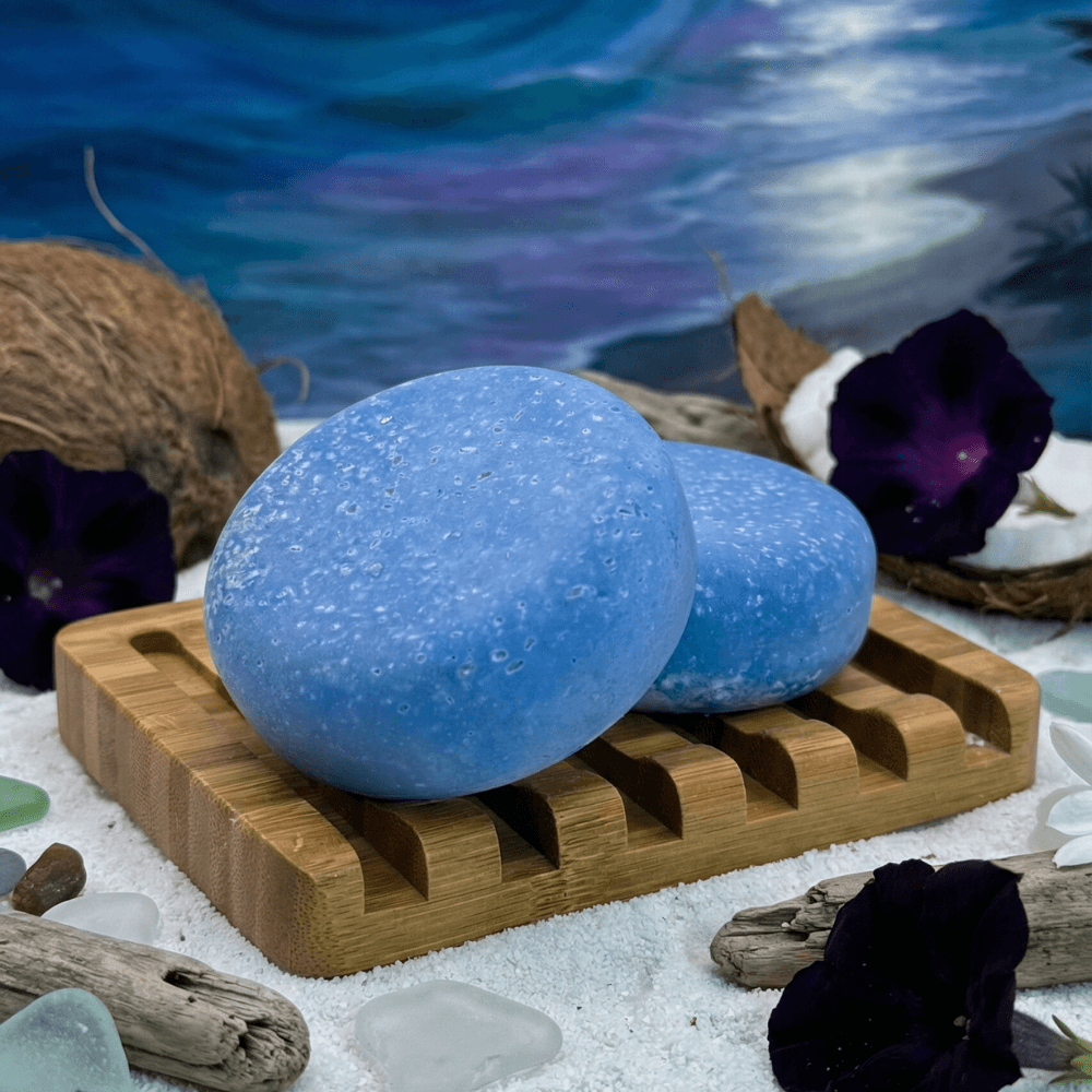 Two oval blue salt scrub bars with crystalline texture sit on a bamboo soap dish surrounded by coconuts, driftwood, purple flowers, and sea glass against a white sand and ocean backdrop.
