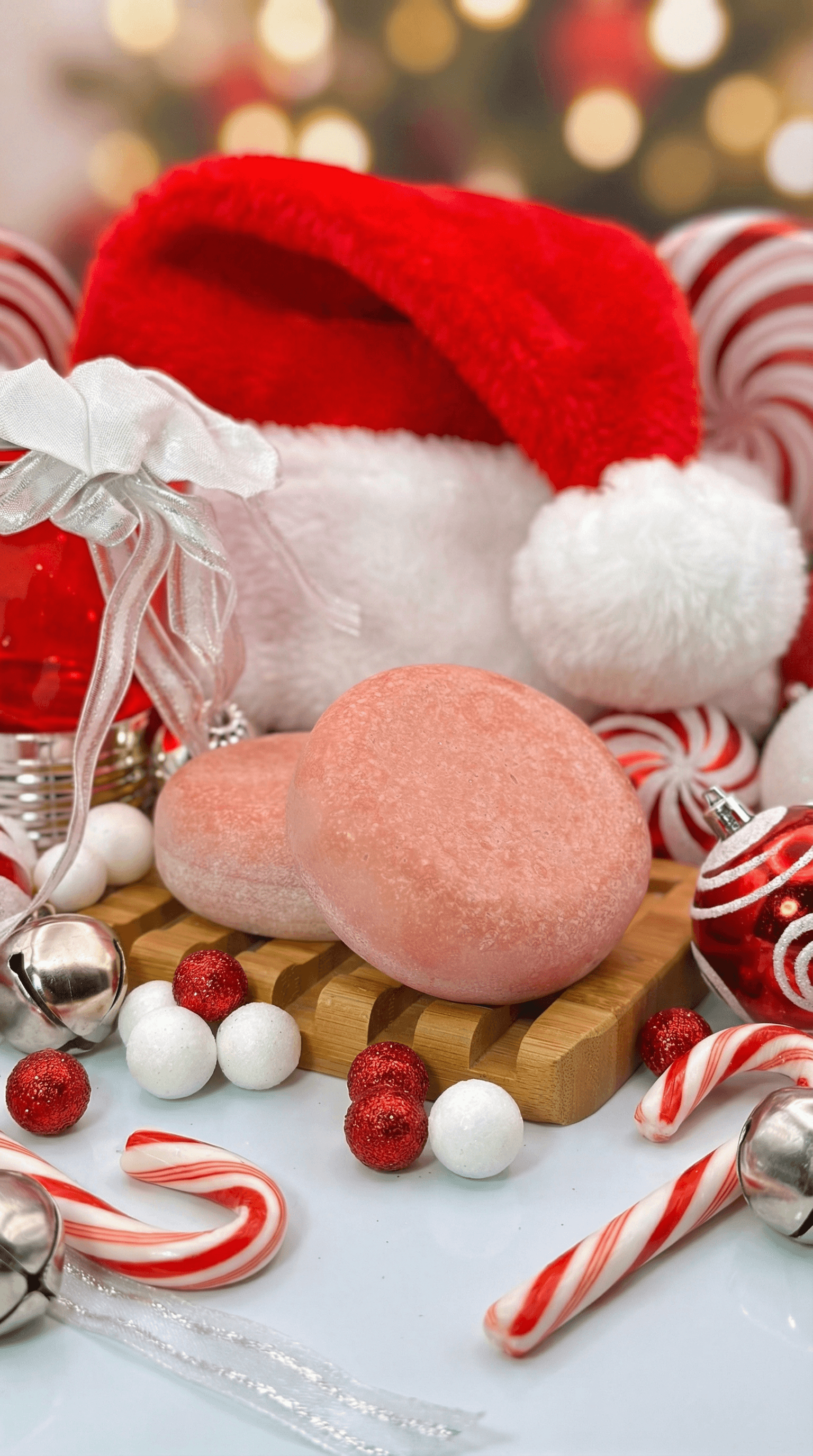 Two pink, sugar-dusted oval sea salt scrub bars on a wooden soap dish, surrounded by red and white Santa hat, candy canes, and festive Christmas ornaments with bokeh lights in background.