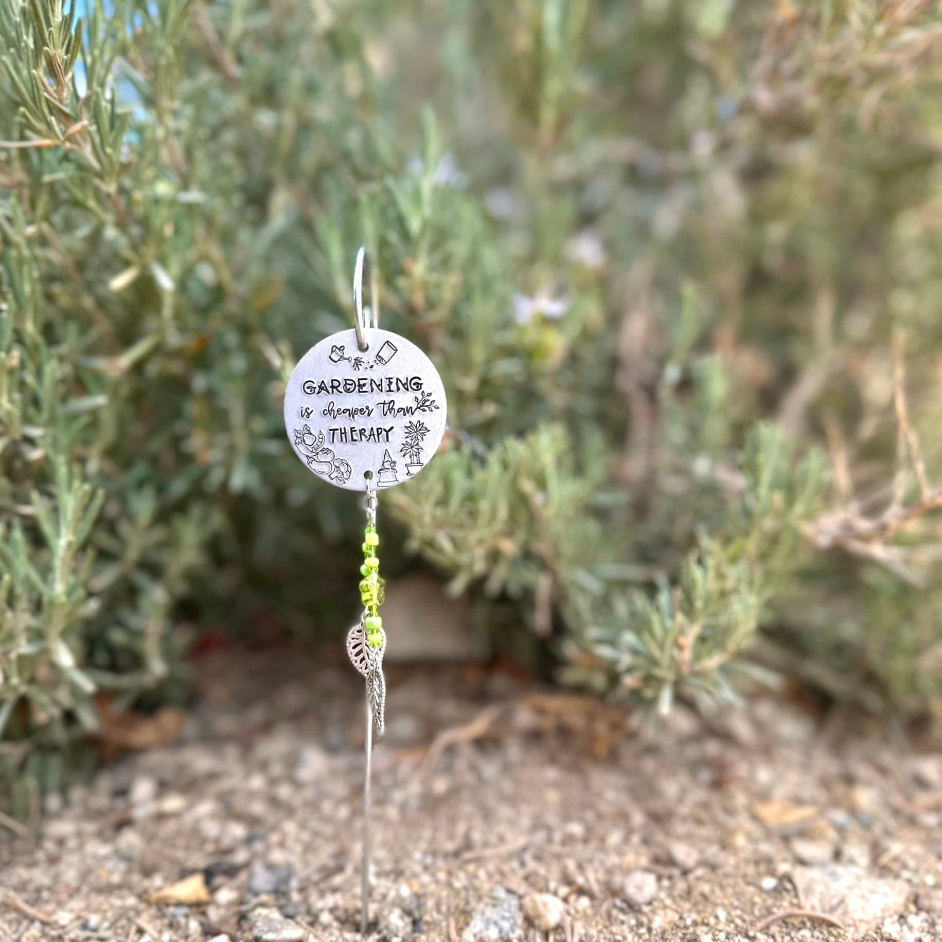 Hand-stamped aluminum garden stake with engraved text reading 'Gardening is cheaper than therapy' on a circular disc, decorated with small gardening icons and green glass beads, photographed outdoors among herbs.