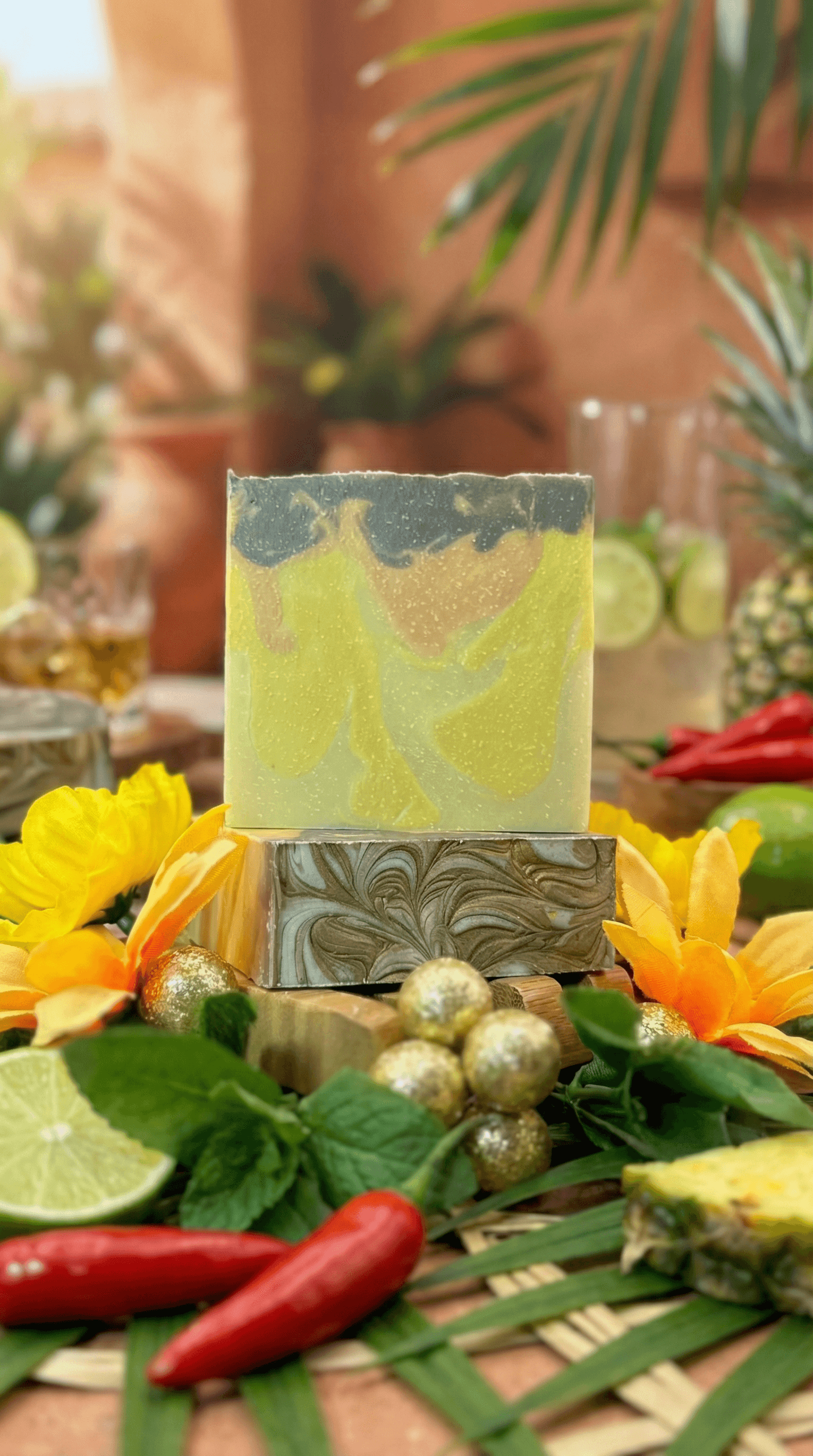 Two stacked rectangular soap bars with swirled yellow, green, and grey marbled pattern, displayed on woven mat surrounded by fresh sunflowers, lime slices, red chilies, mint leaves, and golden berries.