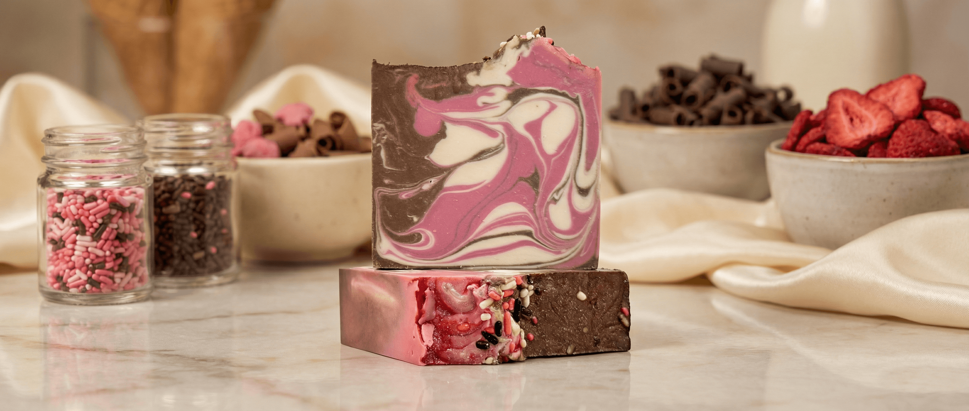 Handcrafted soap bars stacked on marble surface with marbled pink, cream, and brown swirls topped with colorful candy sprinkles. Jars of sprinkles and bowls of ingredients visible in background.