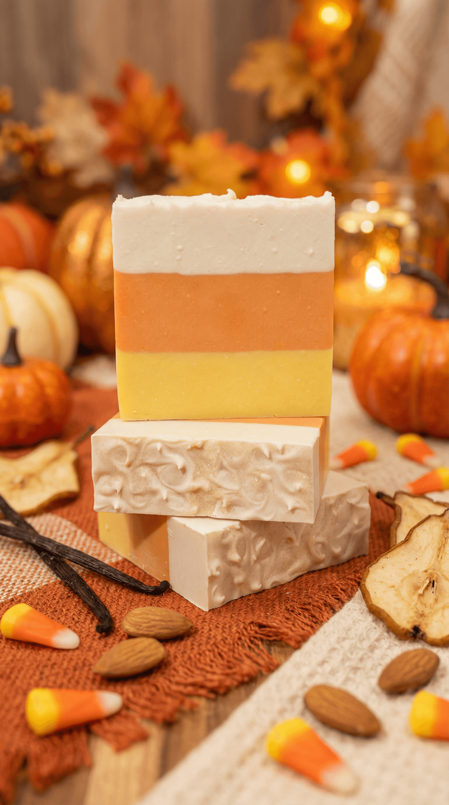 Stack of four handmade soap bars in candy corn colors: white top layer, orange middle, and yellow bottom. Bar in front shows swirled cream texture. Arranged with pumpkins, vanilla beans, almonds, and candy corn on orange cloth.