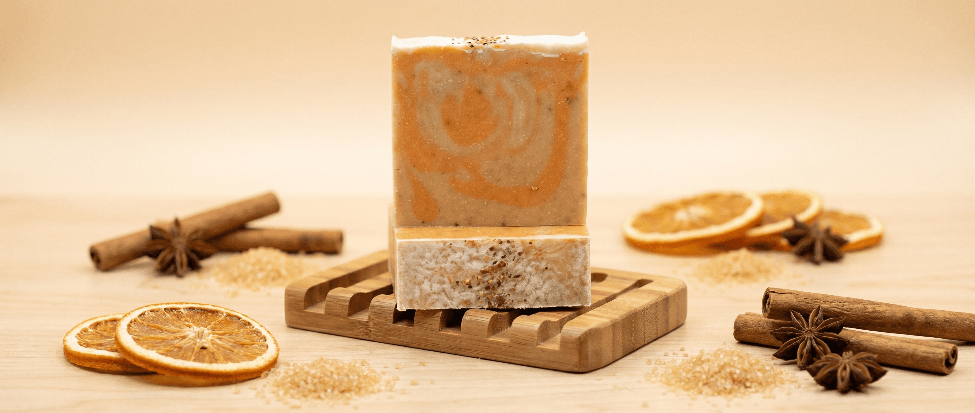 Square orange and cream swirled soap bar with speckled texture on wooden soap dish, surrounded by dried orange slices, cinnamon sticks, and star anise on neutral background.
