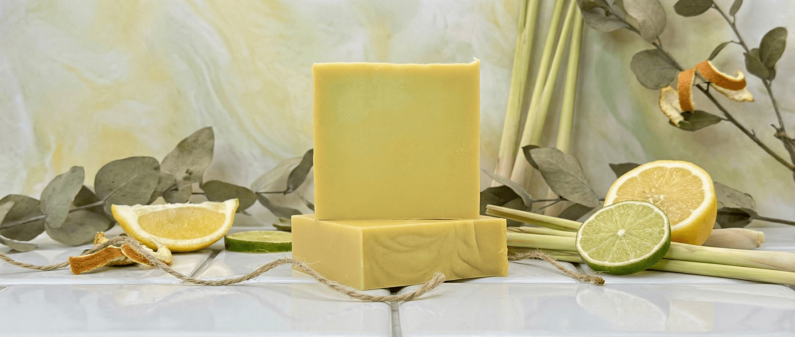 Pale yellow soap bars with smooth matte finish arranged with fresh lemongrass stalks, eucalyptus sprigs, and halved citrus fruits on white marble surface.