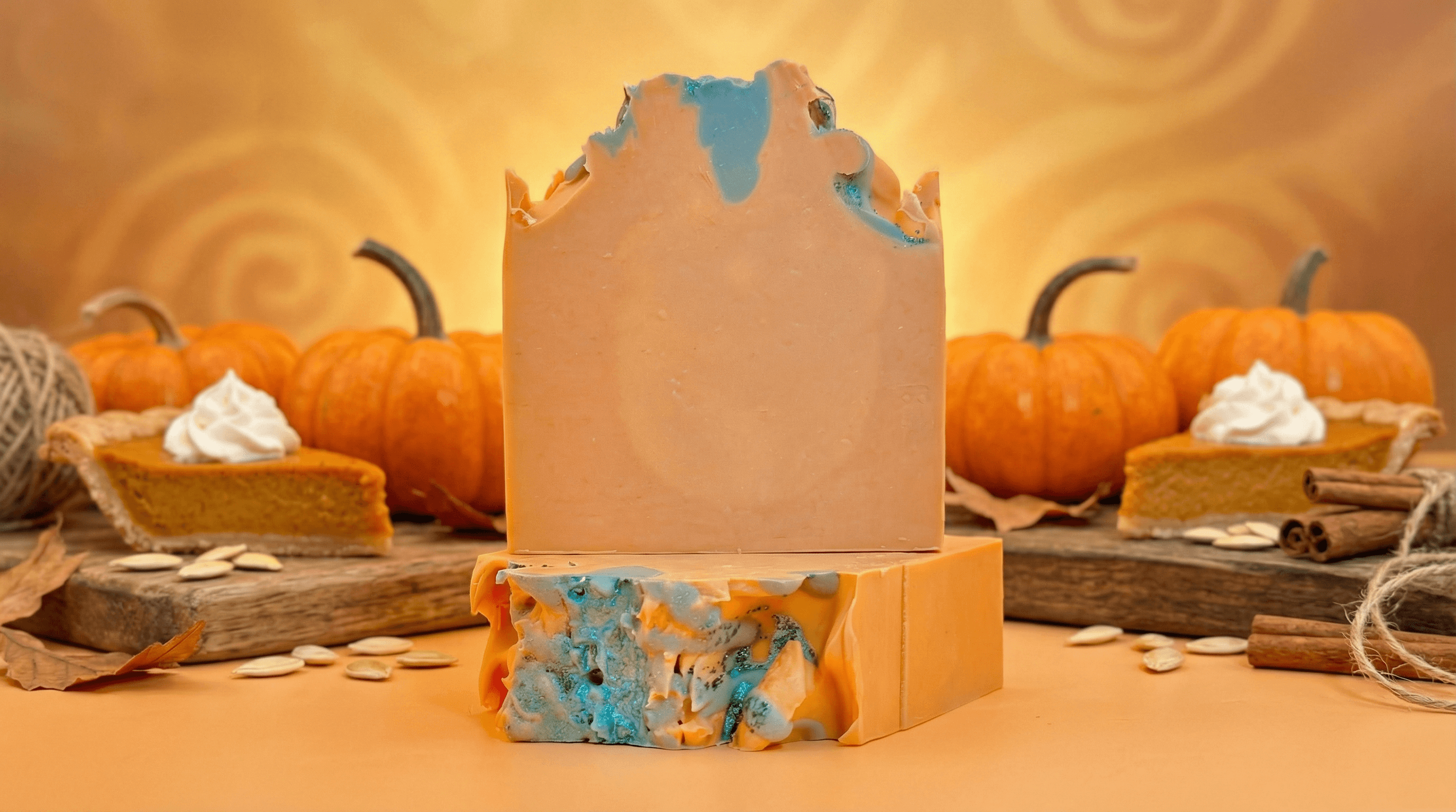 Handcrafted soap bar with warm orange base and swirled blue-green marbled top, displayed standing and sliced against autumn setting with pumpkins, pumpkin pie slices, and cinnamon sticks.