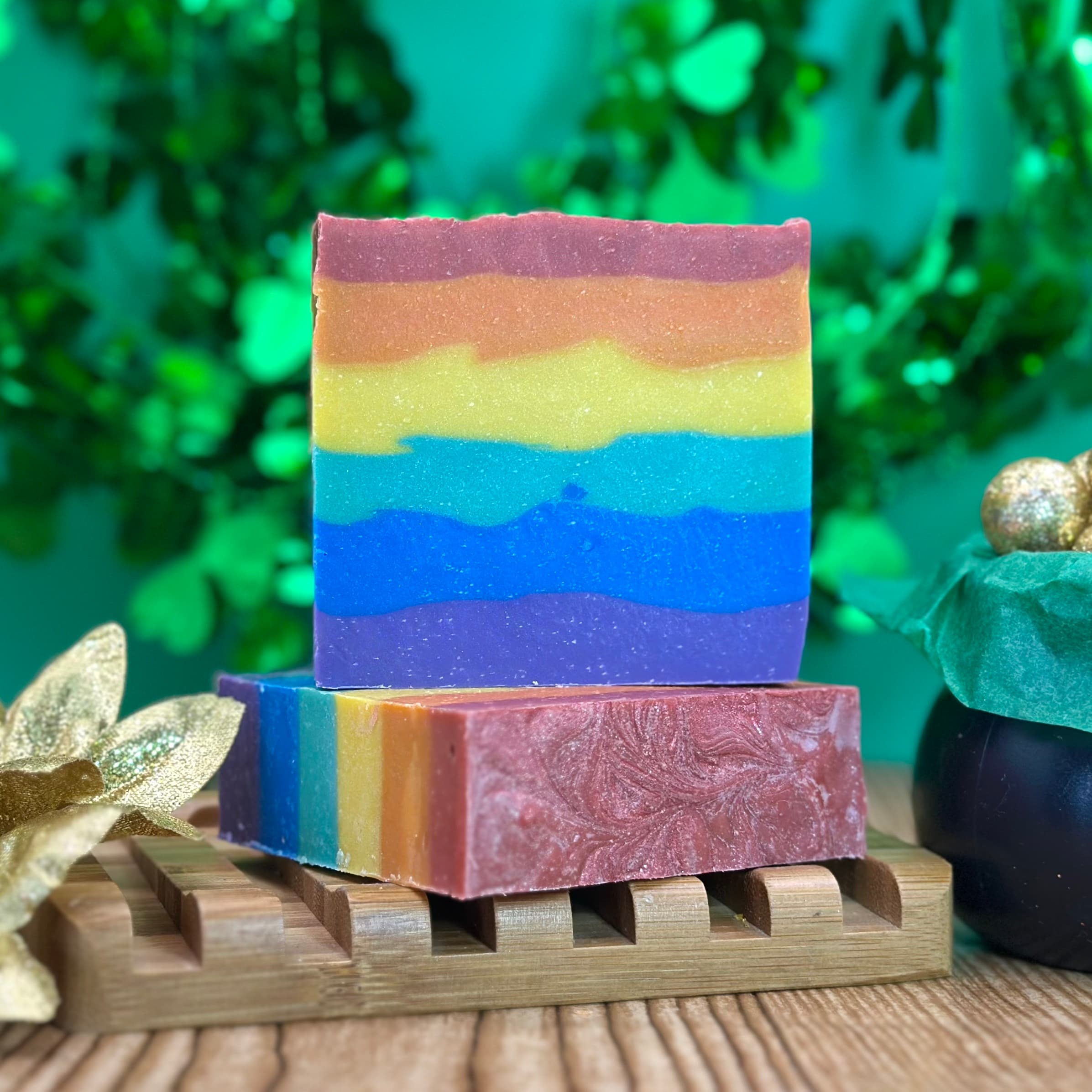 Two rainbow-striped rectangular soap bars stacked on a wooden soap dish, displaying vibrant horizontal layers from purple through blue, yellow, orange, to mauve, with a shimmer finish.