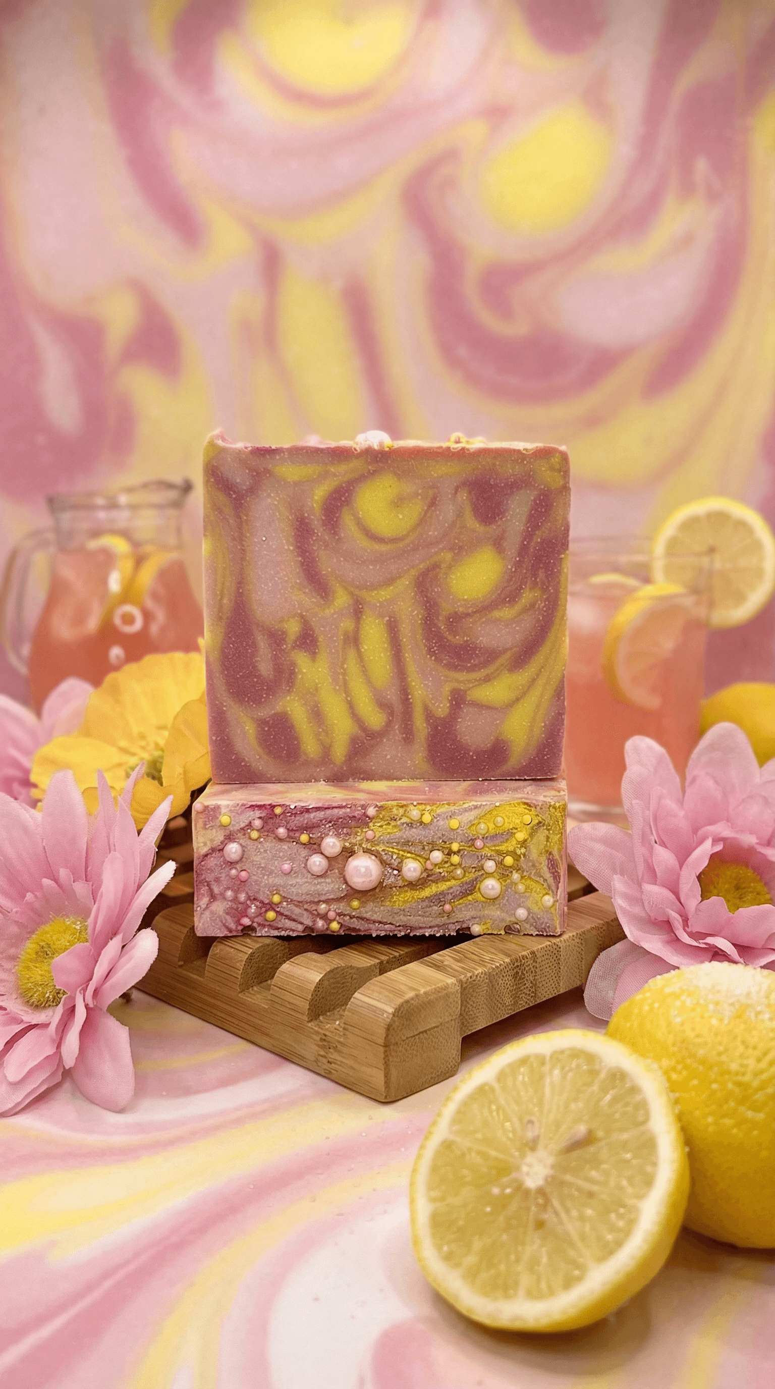 Two stacked bars of handmade soap with pink and yellow marbled swirls on top, embedded pearls and sparkles on bottom half, displayed on wooden soap dish with pink flowers and fresh lemons.