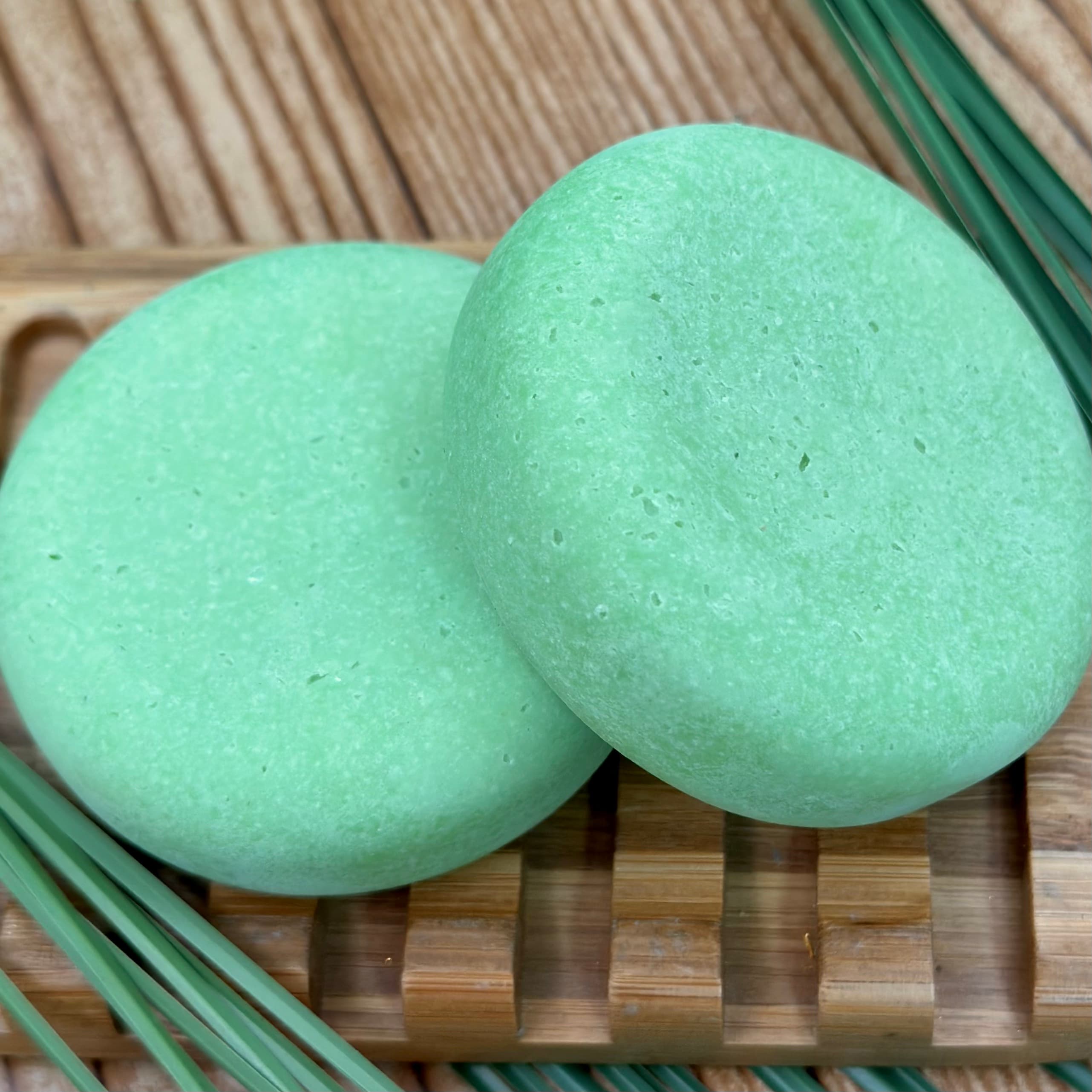 Two pale mint green round bars with visible flecks throughout, resting on a wooden soap dish with bamboo leaves as accents against a textured neutral background.