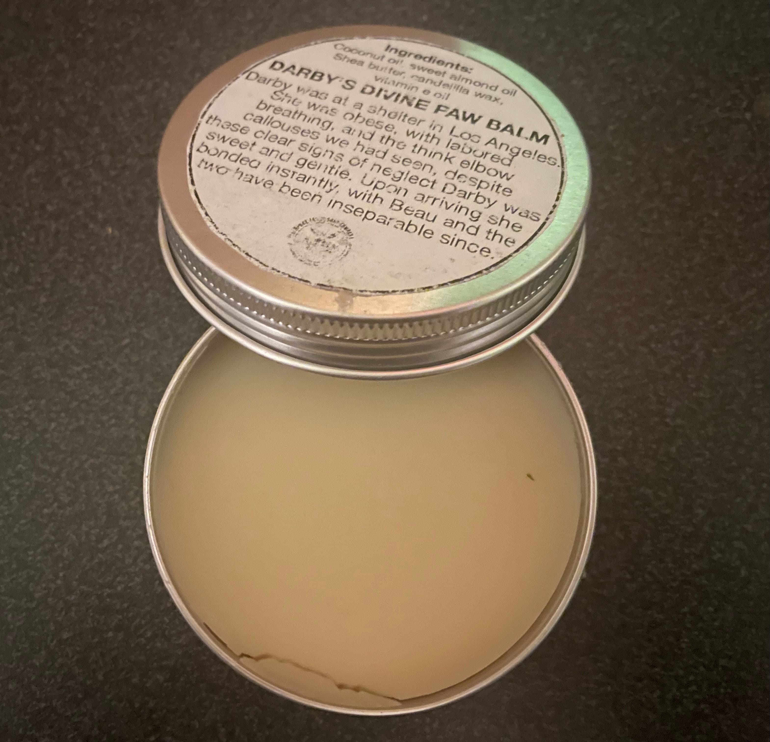Open tin of pale cream-colored paw balm with smooth surface. Metal container with printed lid showing product information resting beside the open tin.